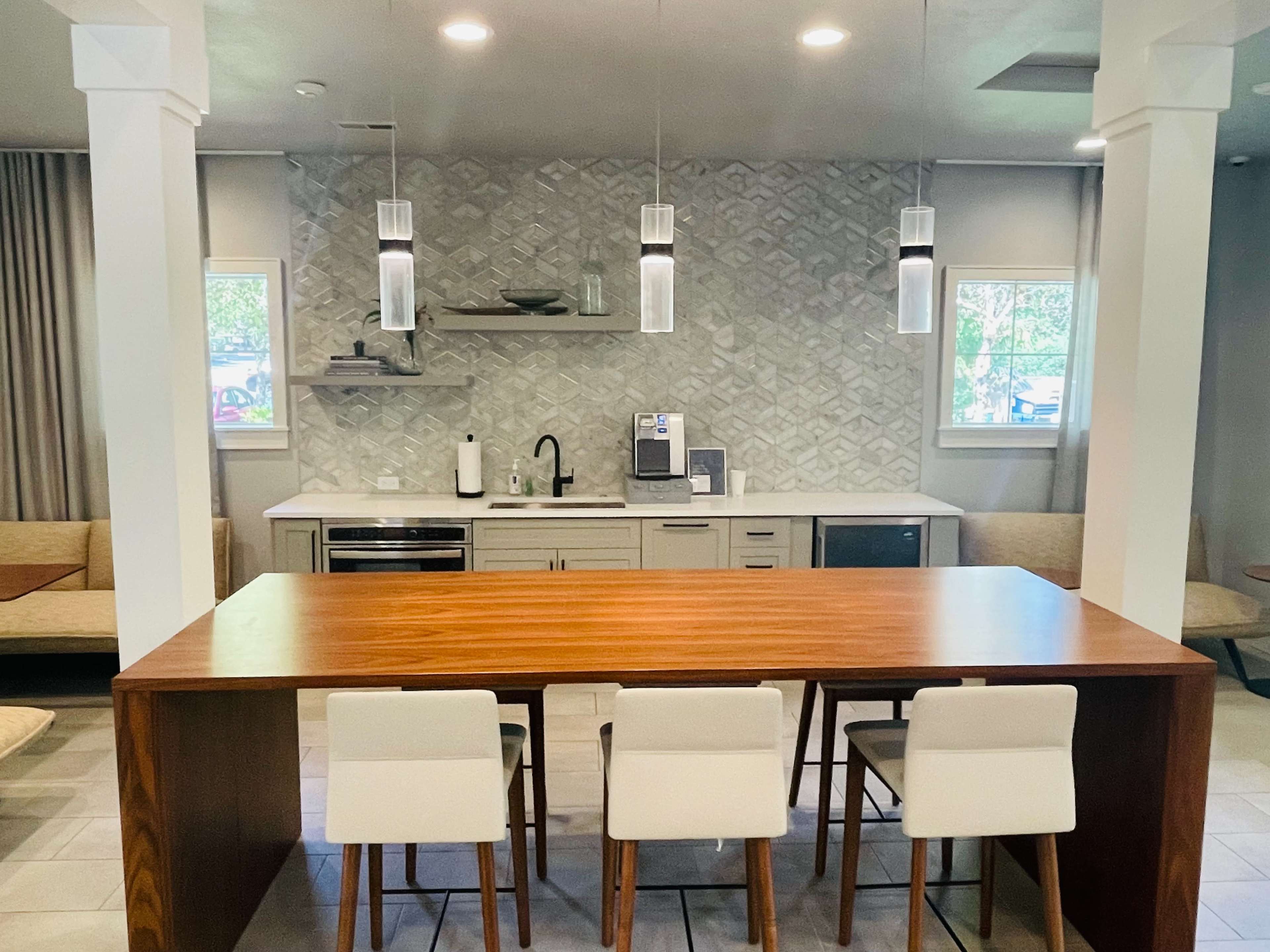 A modern kitchen area with a wooden table and white bar stools, featuring a herringbone-patterned backsplash and appliances against a light-colored wall.