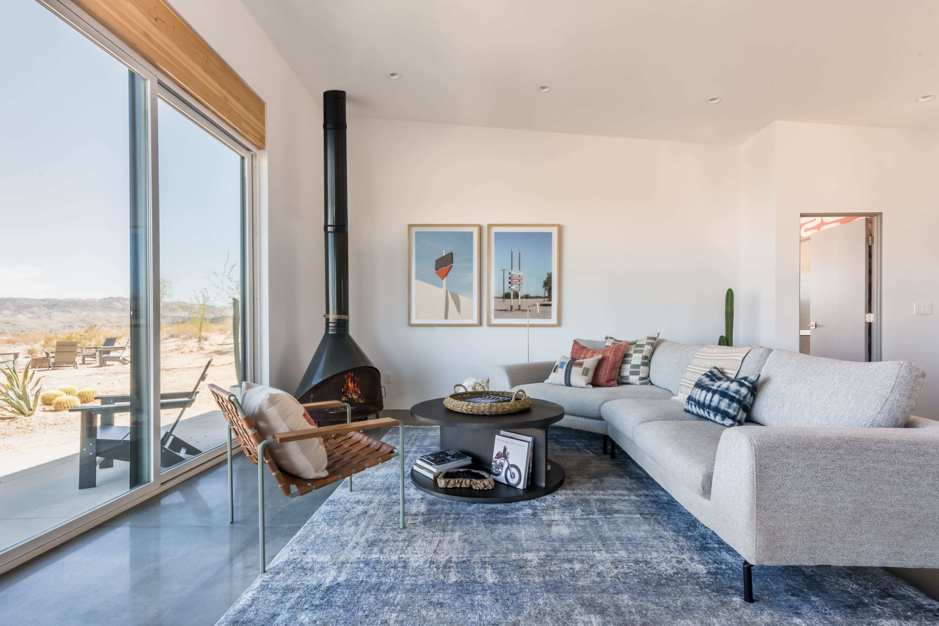 A modern living room features a light gray sectional sofa, a round coffee table, and two framed artworks, with large windows revealing a desert landscape outside.