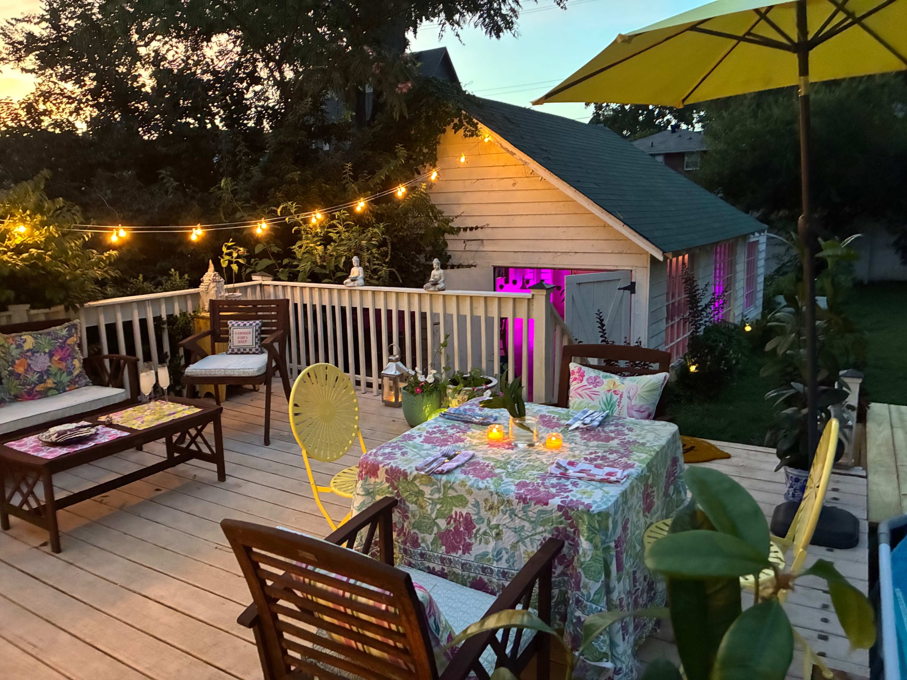 The image shows a warmly lit outdoor patio with wooden decking, colorful furniture, and a charming shed in the background.