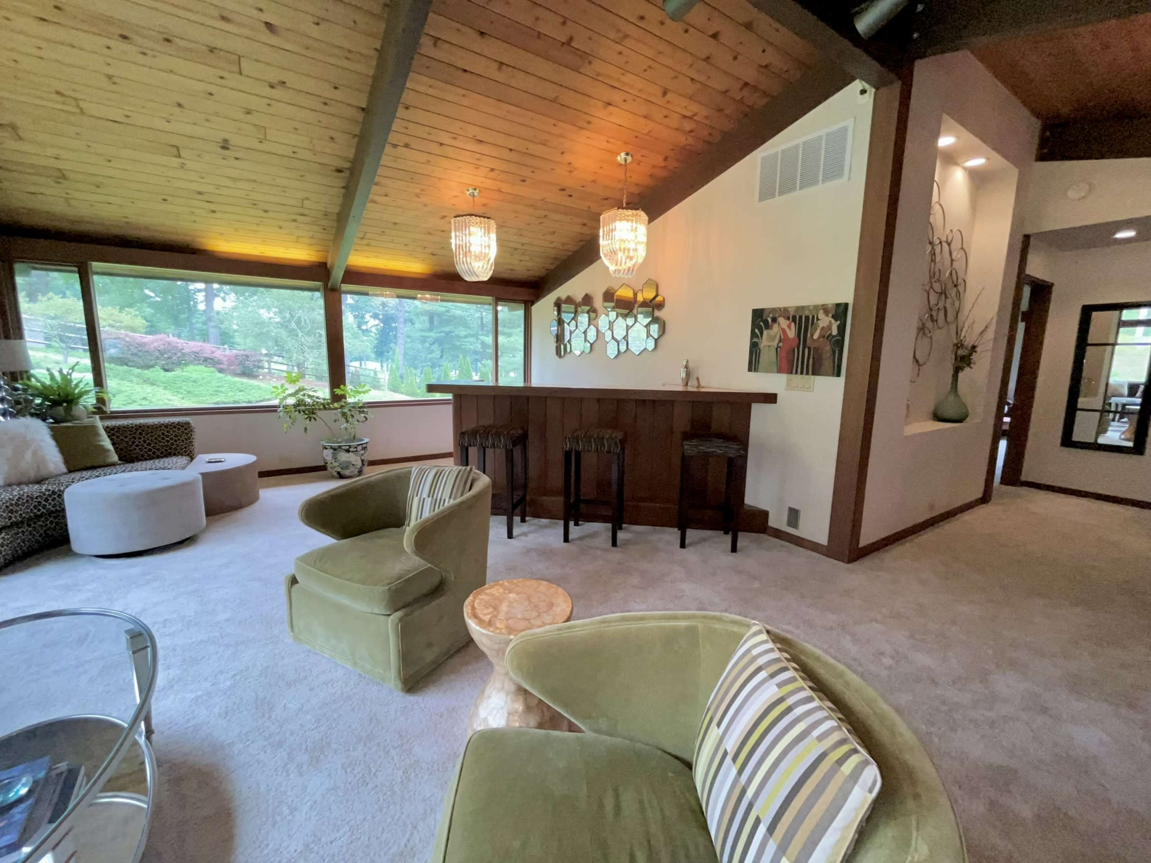 The image shows a spacious living room with a bar area, featuring a wood-paneled ceiling and large windows overlooking greenery.