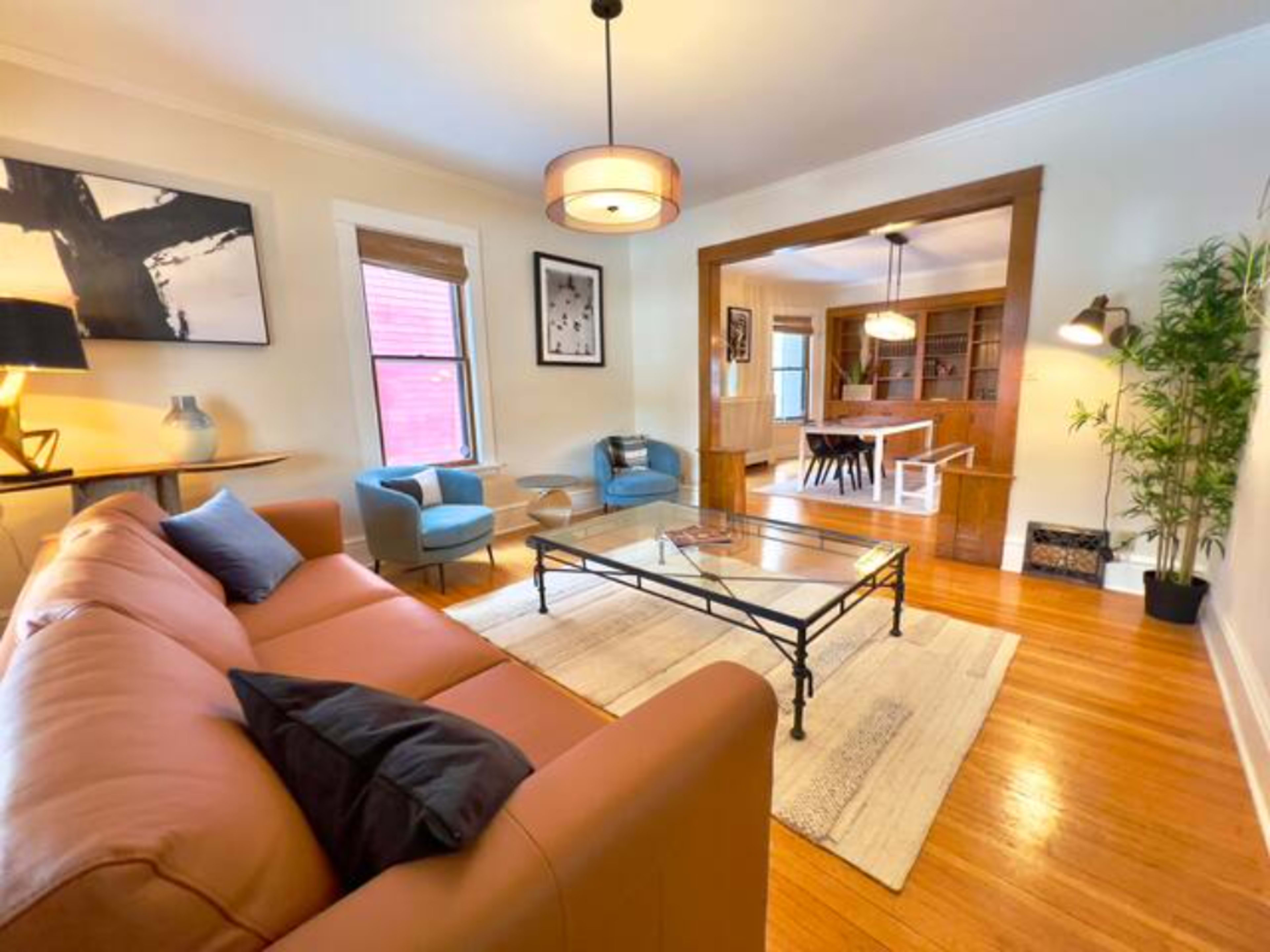 A living room features a brown sofa, glass coffee table, and two blue armchairs, with a view into a dining area through an open archway.