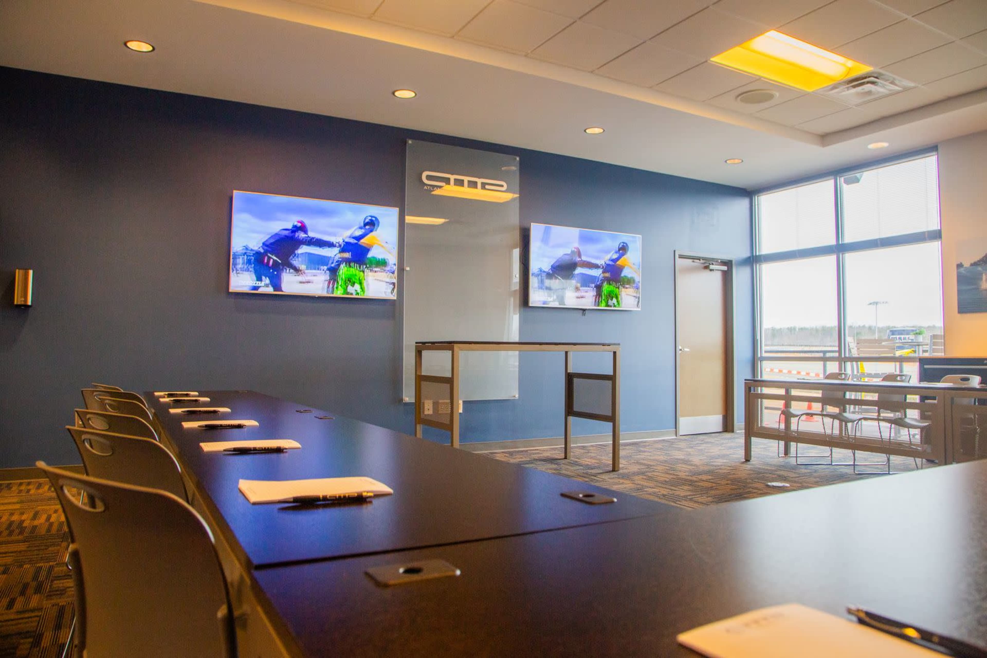 The image shows a modern conference room with two large screens mounted on the wall, a long table with seating, and a glass door leading outside.