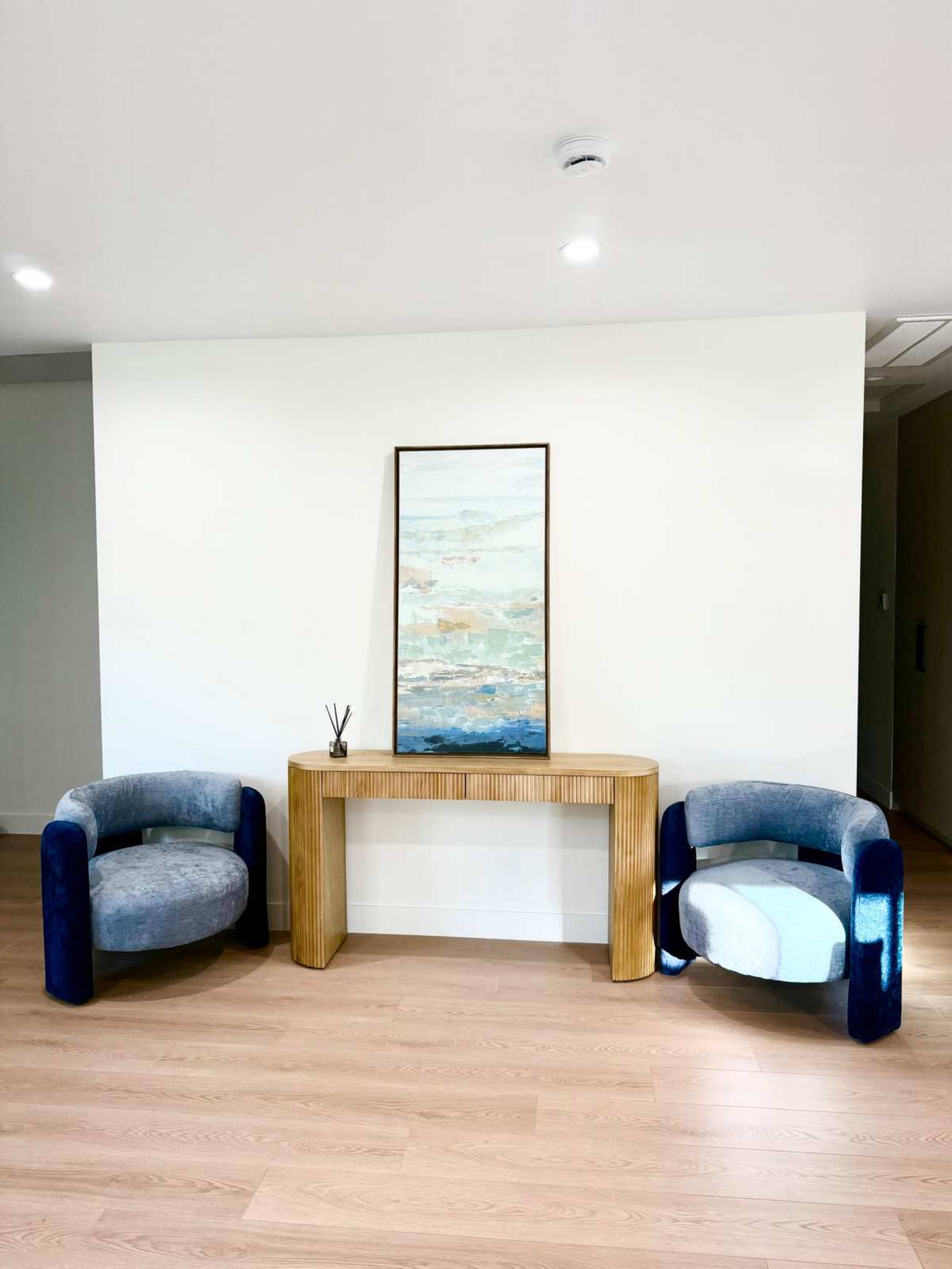 Two upholstered chairs with curved backs flank a wooden console table, which supports a framed abstract painting, in a minimalist interior space with light wooden flooring.