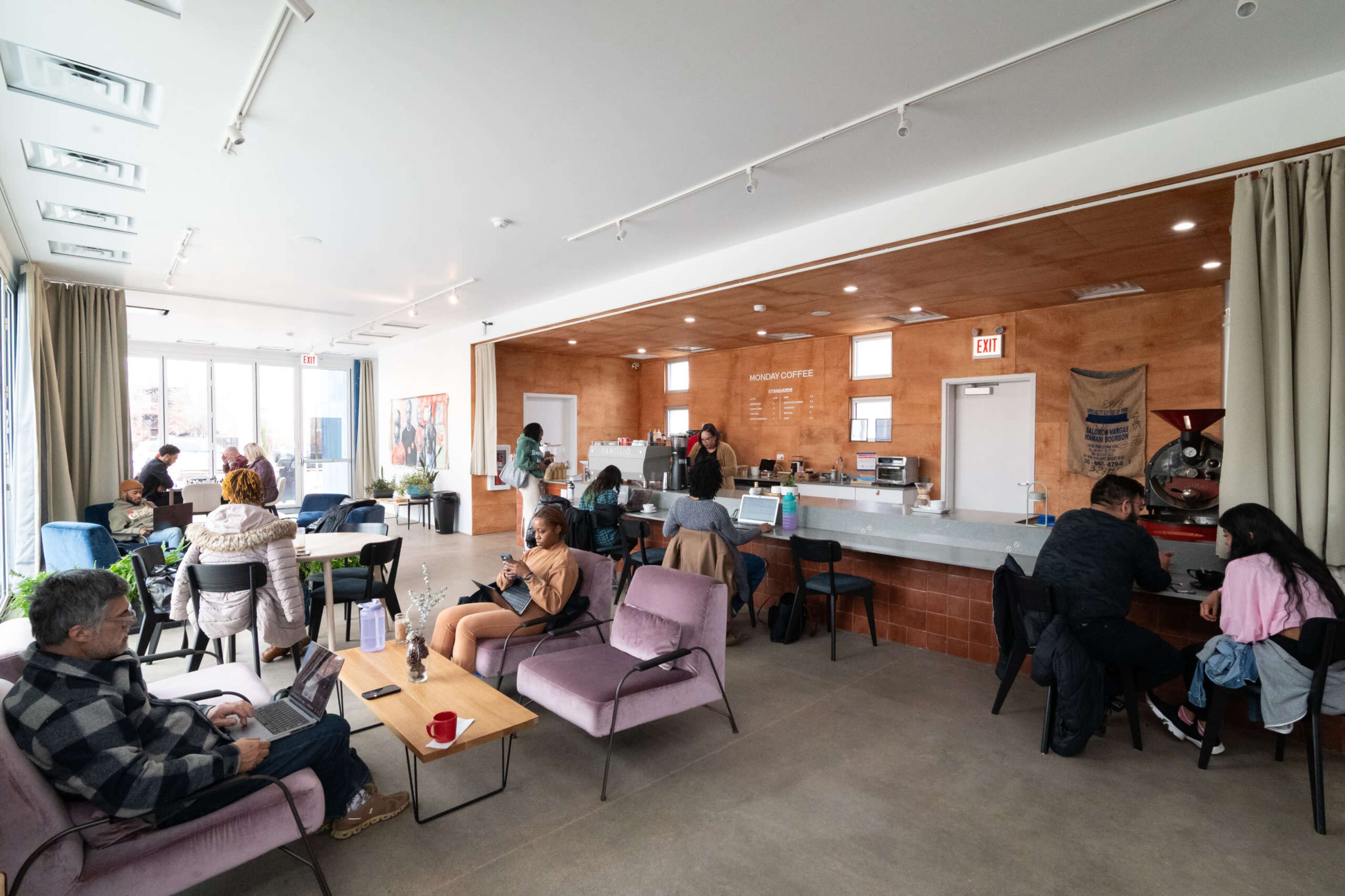 The image showcases a modern café interior with patrons seated on chairs and couches, working on laptops, and enjoying their drinks at a counter.