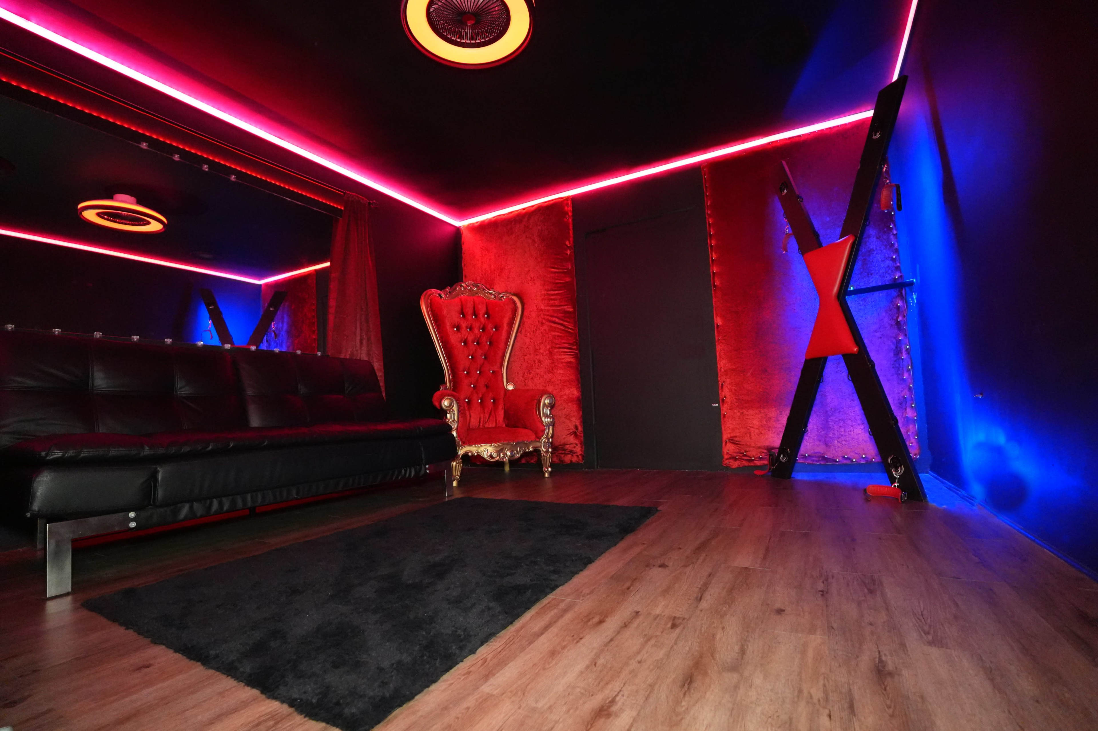 The image shows a dark room with red and blue neon lights, featuring a black sofa, a red velvet chair, and a textured wall.