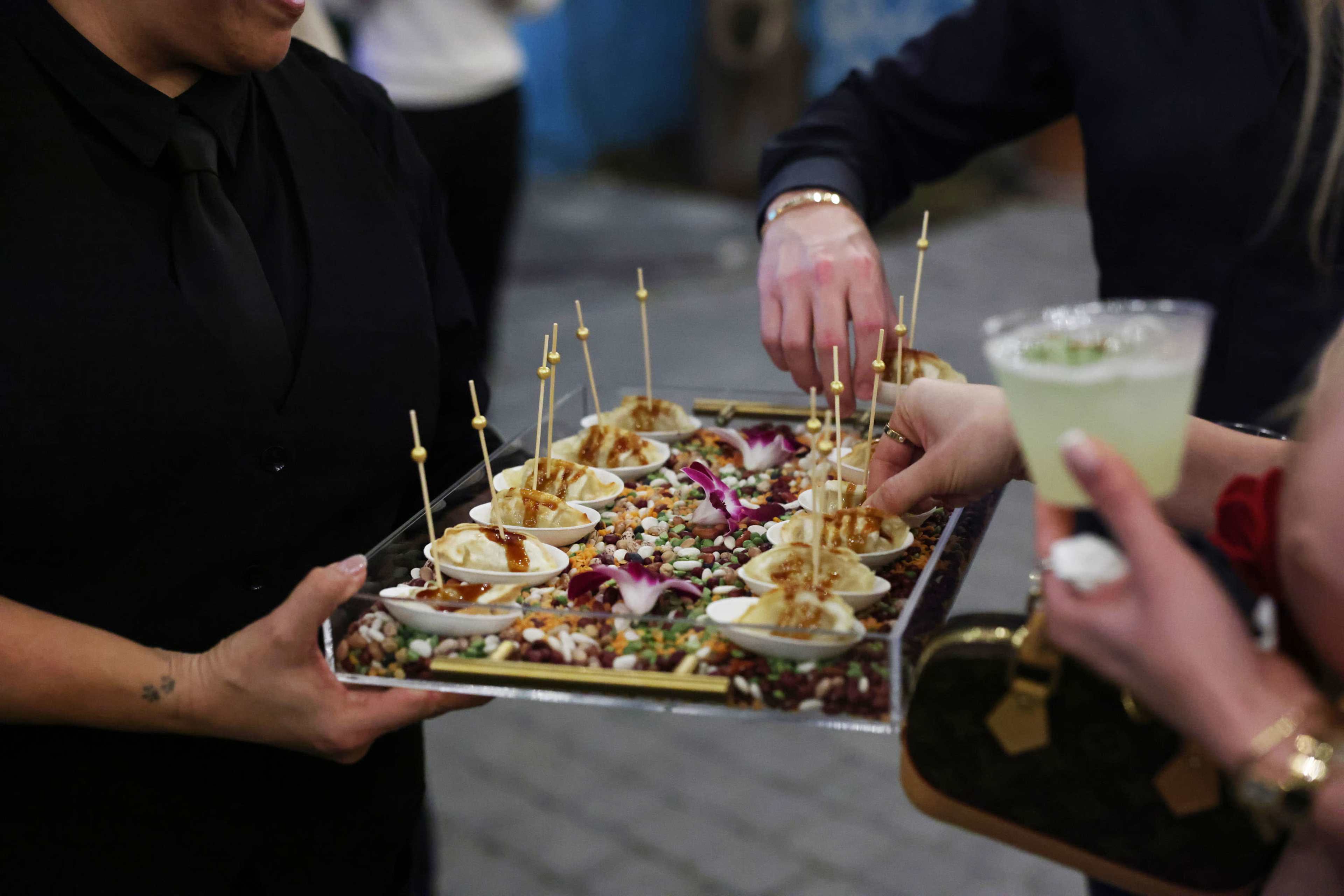 A server offers a tray of appetizers featuring small dishes garnished with edible flowers and decorative toppings, while guests reach for the food and hold beverages.