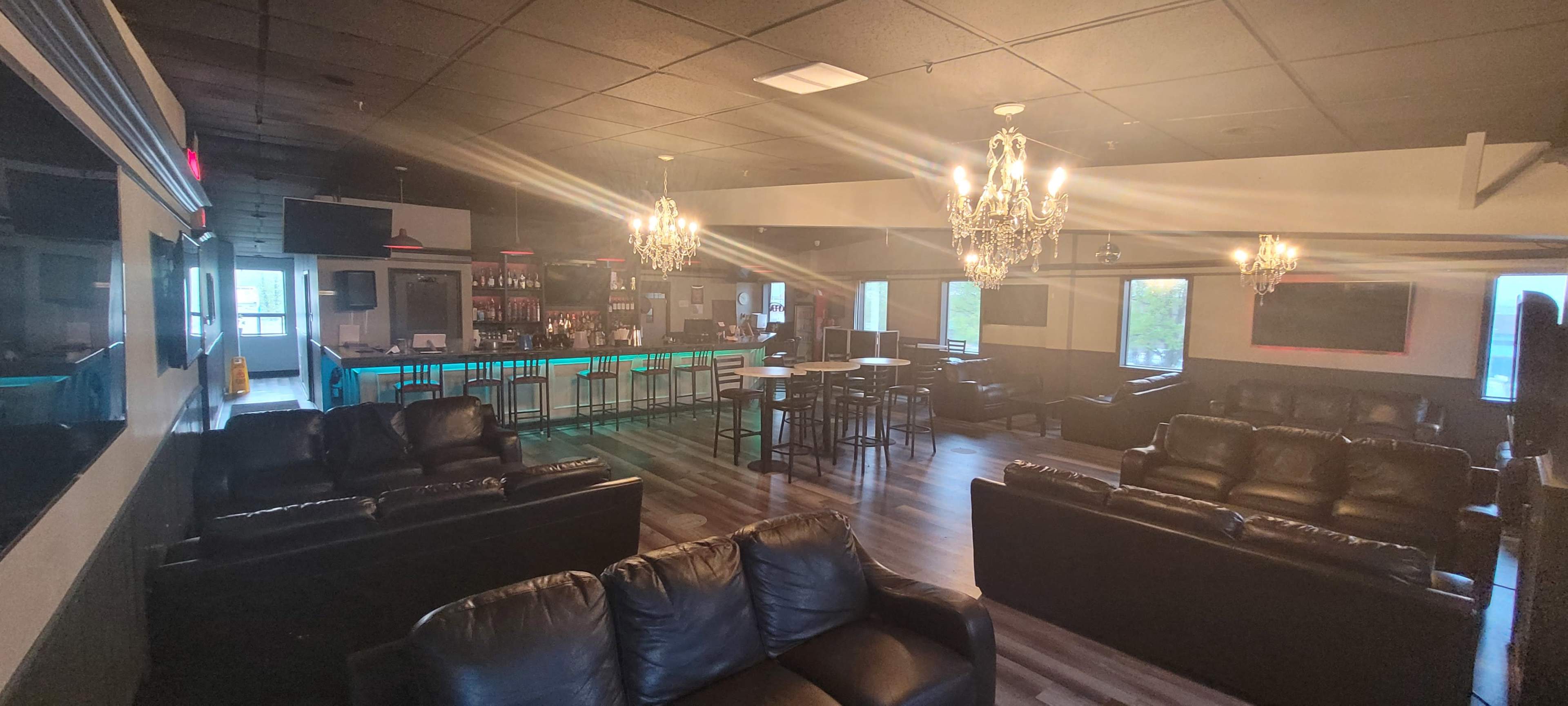 A spacious lounge area with black leather sofas, a bar with blue lighting, and chandeliers hanging from the ceiling.