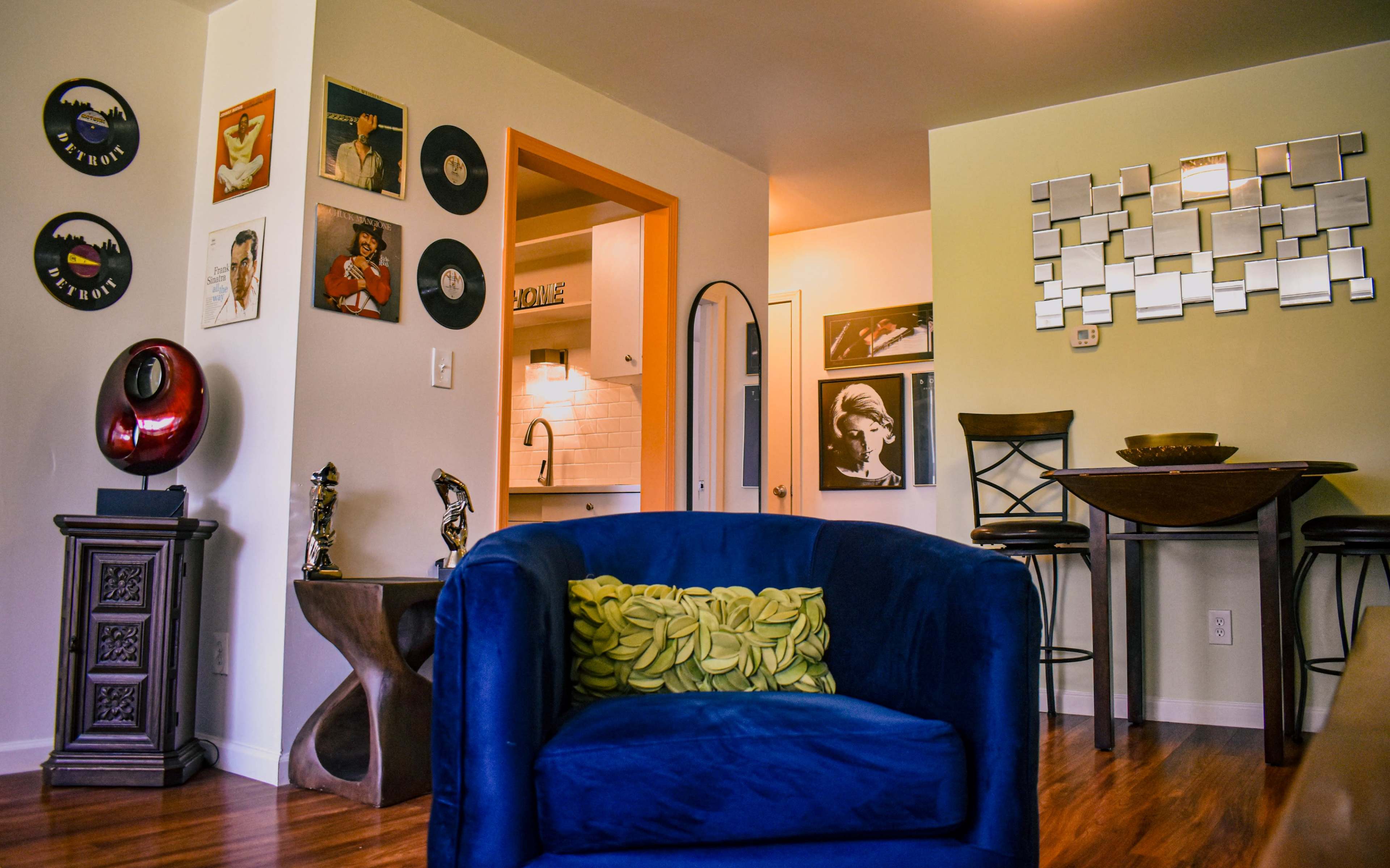 The image shows a cozy living space featuring a blue armchair with yellow accent pillows, wall art including framed records and portraits, and a kitchen area visible in the background.