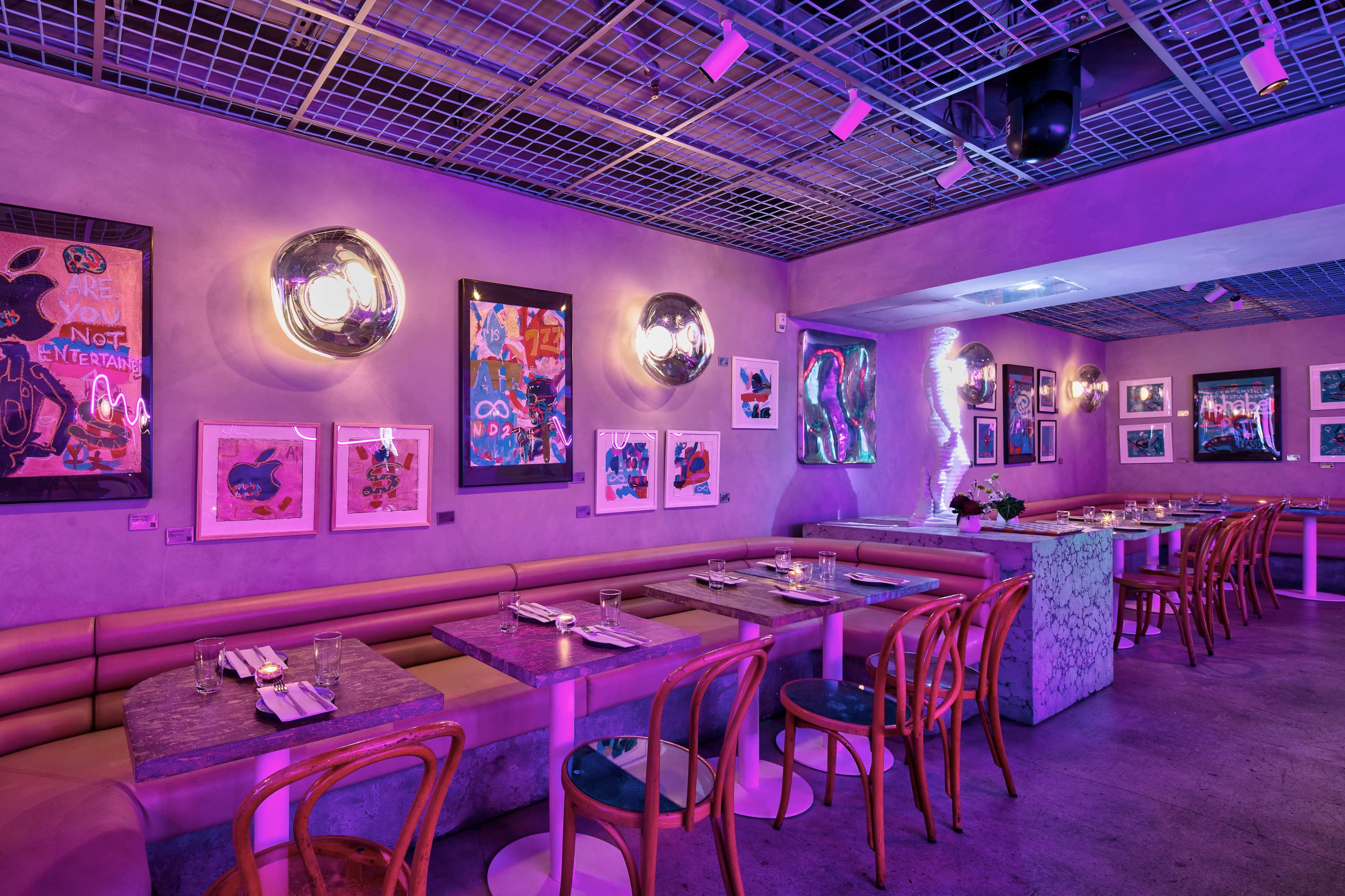 The image shows a modern restaurant interior with purple lighting, featuring artwork on the walls and long tables with seating along the sides.