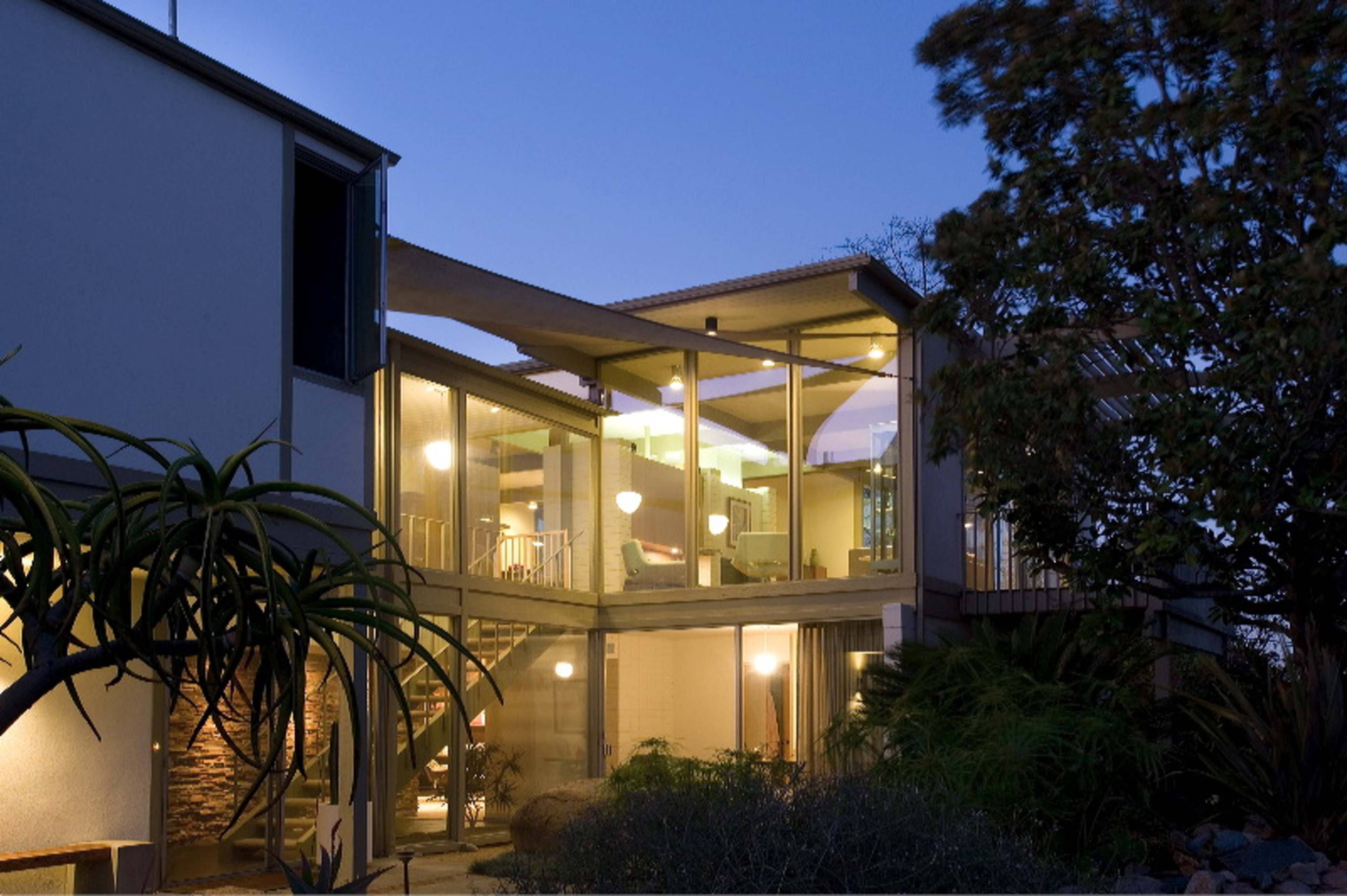 A modern house with large glass windows and a curved design, illuminated at dusk with surrounding greenery.