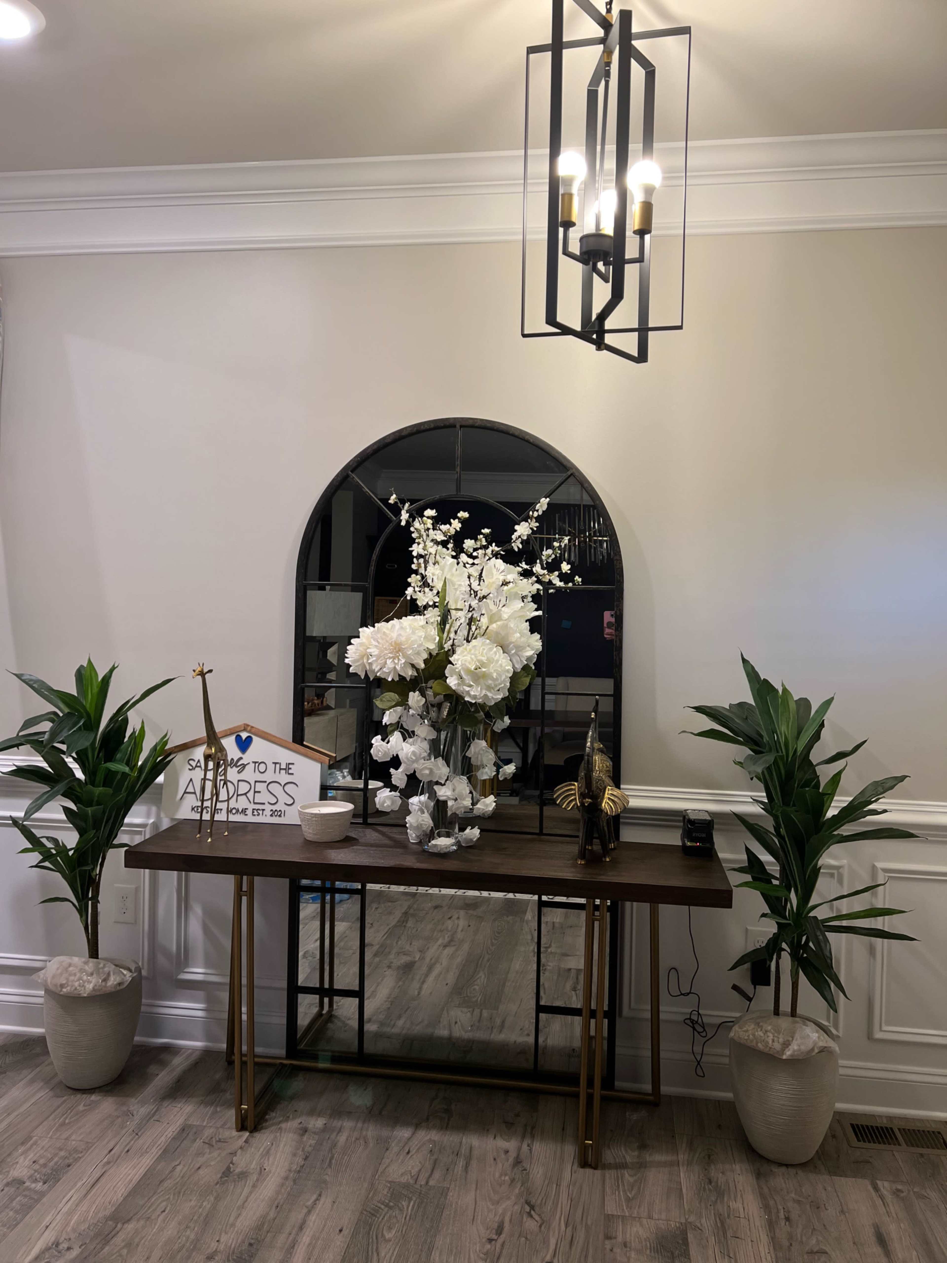 A console table with a large mirror above it is adorned with decorative flowers, a small statue, and potted plants on either side, all under a pendant light fixture.