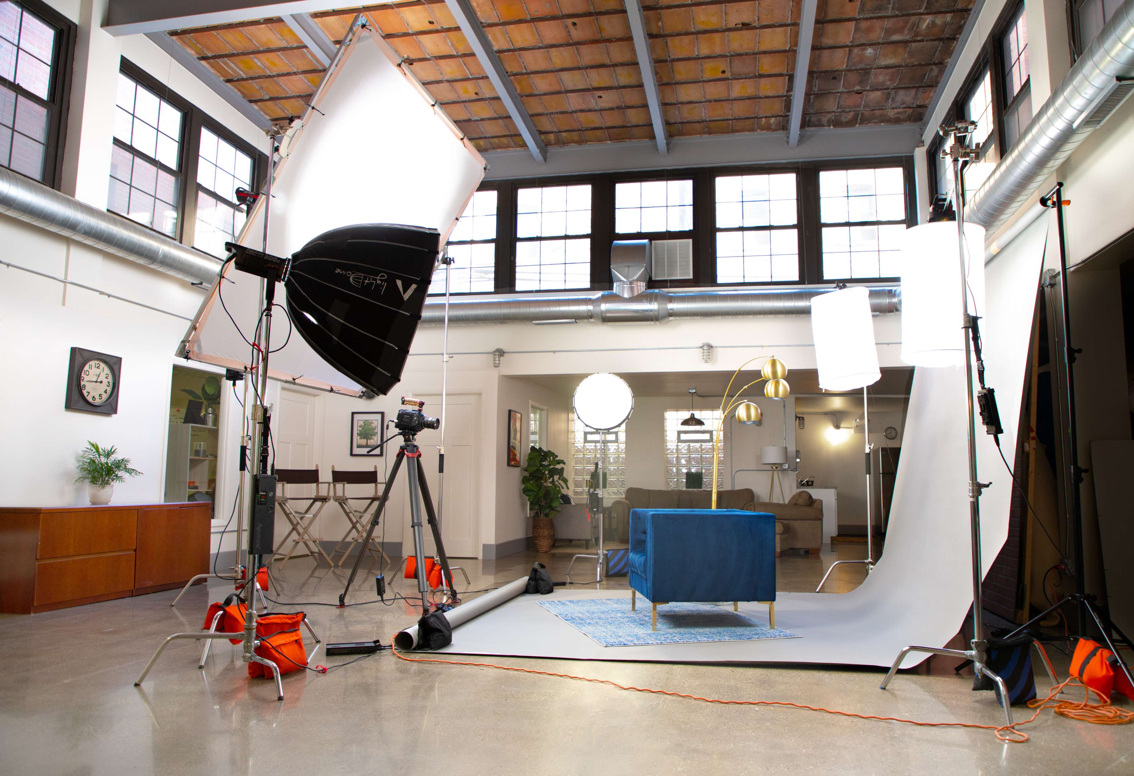 A photography studio setup with lighting equipment, a blue couch, a backdrop, and various props, all arranged in a spacious, well-lit room.