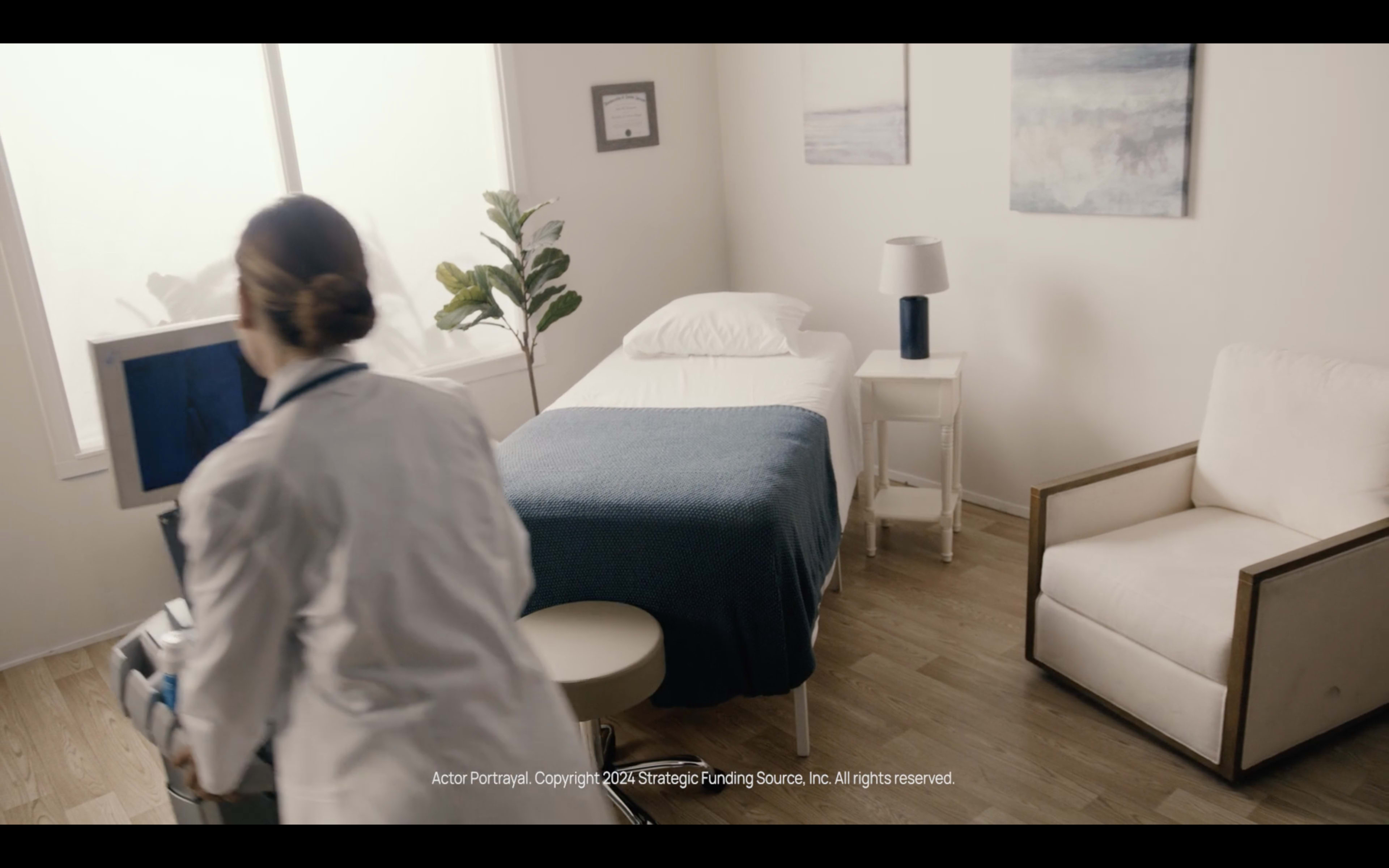 A doctor in a white coat is pushing a medical cart in a well-lit hospital room featuring a single bed, a chair, and a plant by the window.