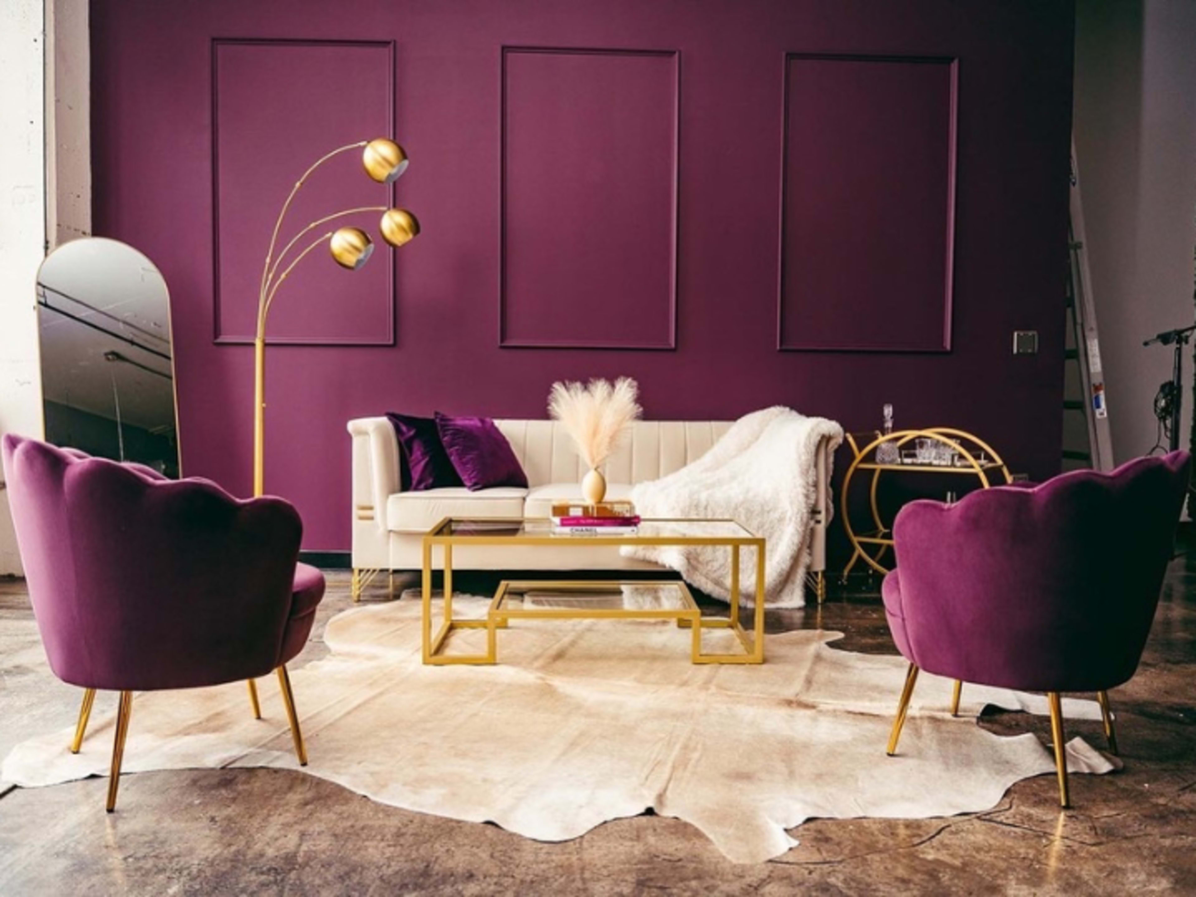 The image shows a modern living room featuring a white sofa, two purple chairs, a gold floor lamp, and a coffee table on a cowhide rug, all set against a deep purple wall.