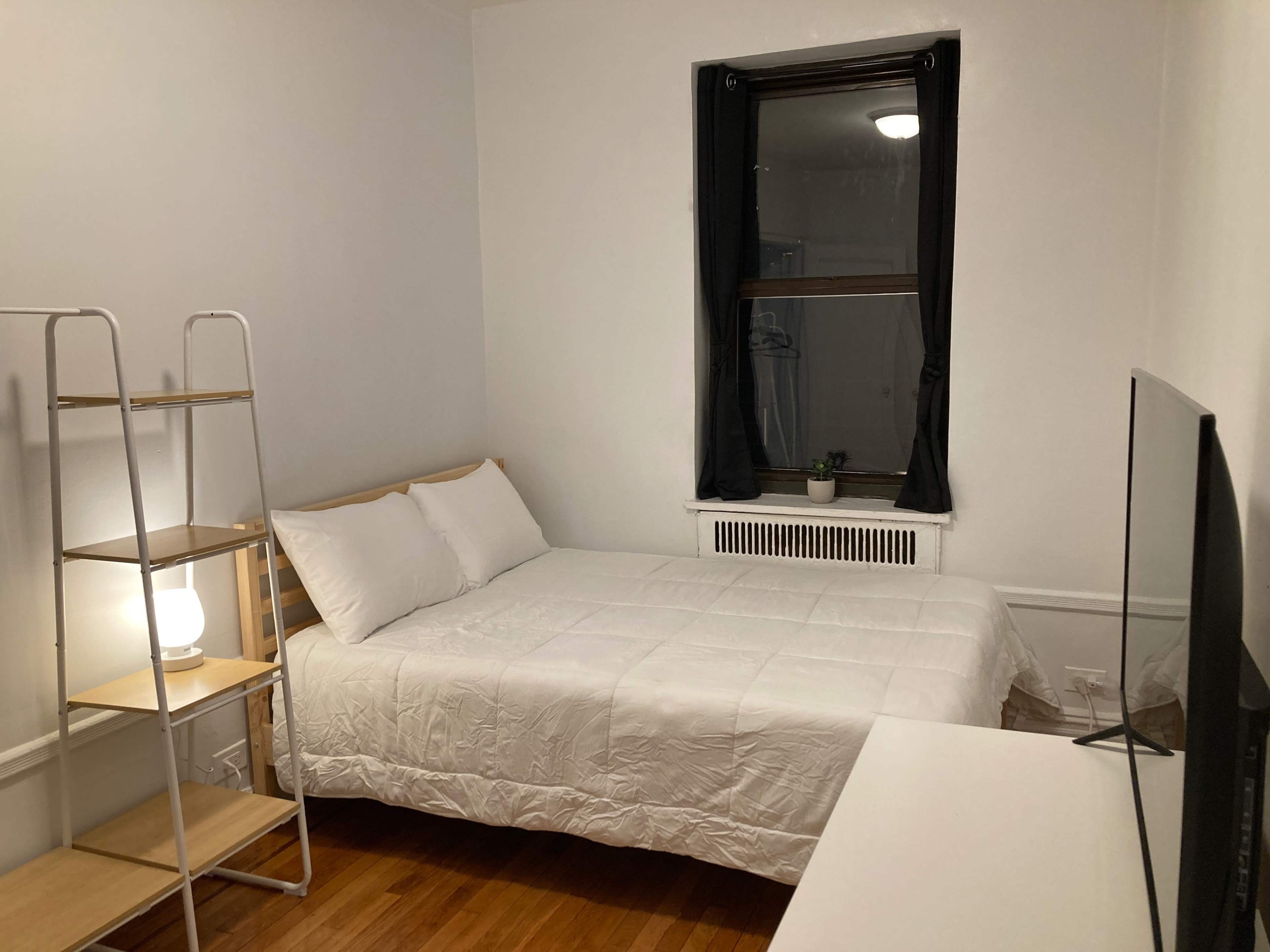 A simple bedroom with a bed, a TV, a small shelf, and a window with curtains.