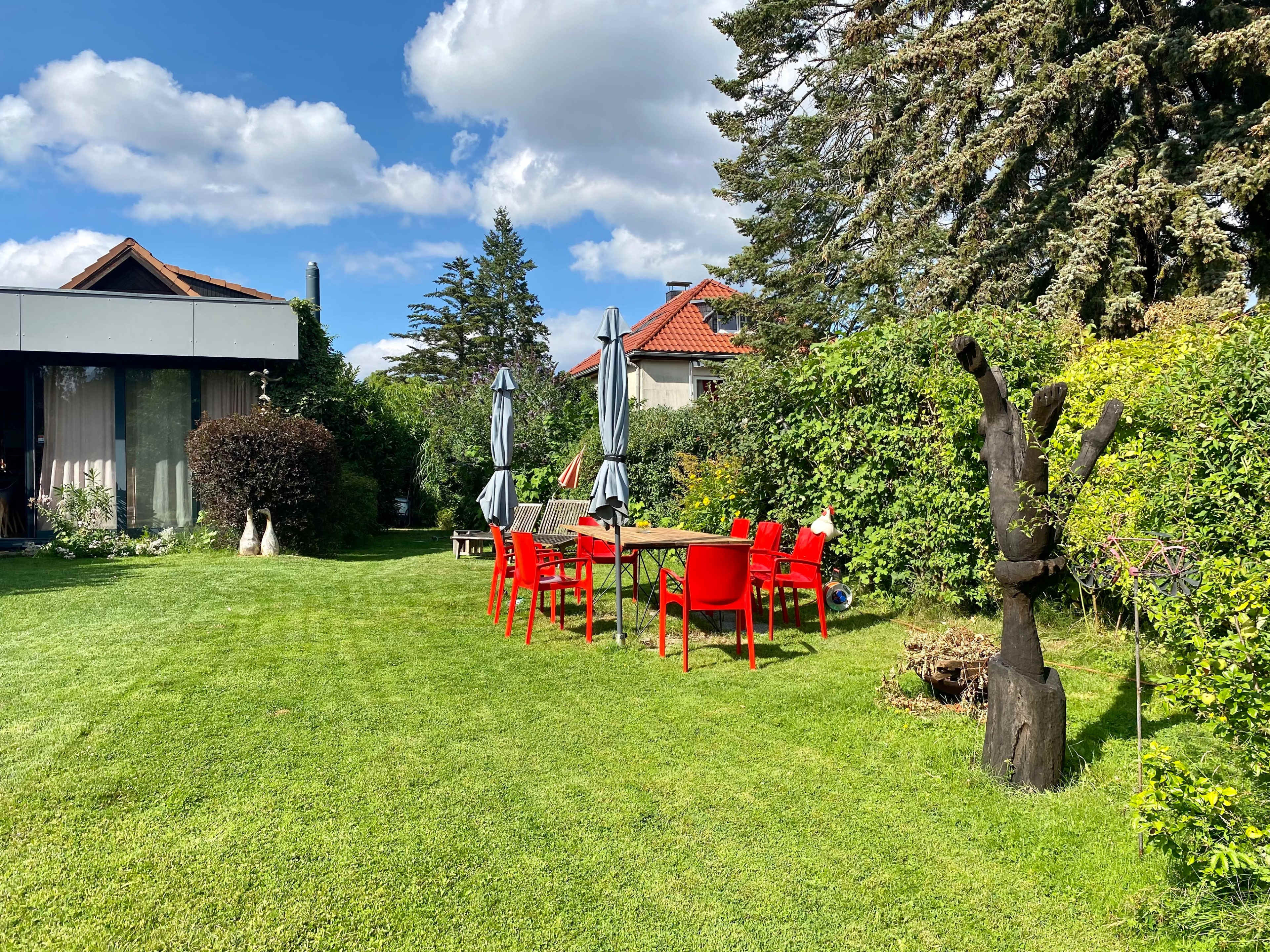 A bright green lawn features a red outdoor dining set surrounded by trees and shrubs under a partly cloudy sky.