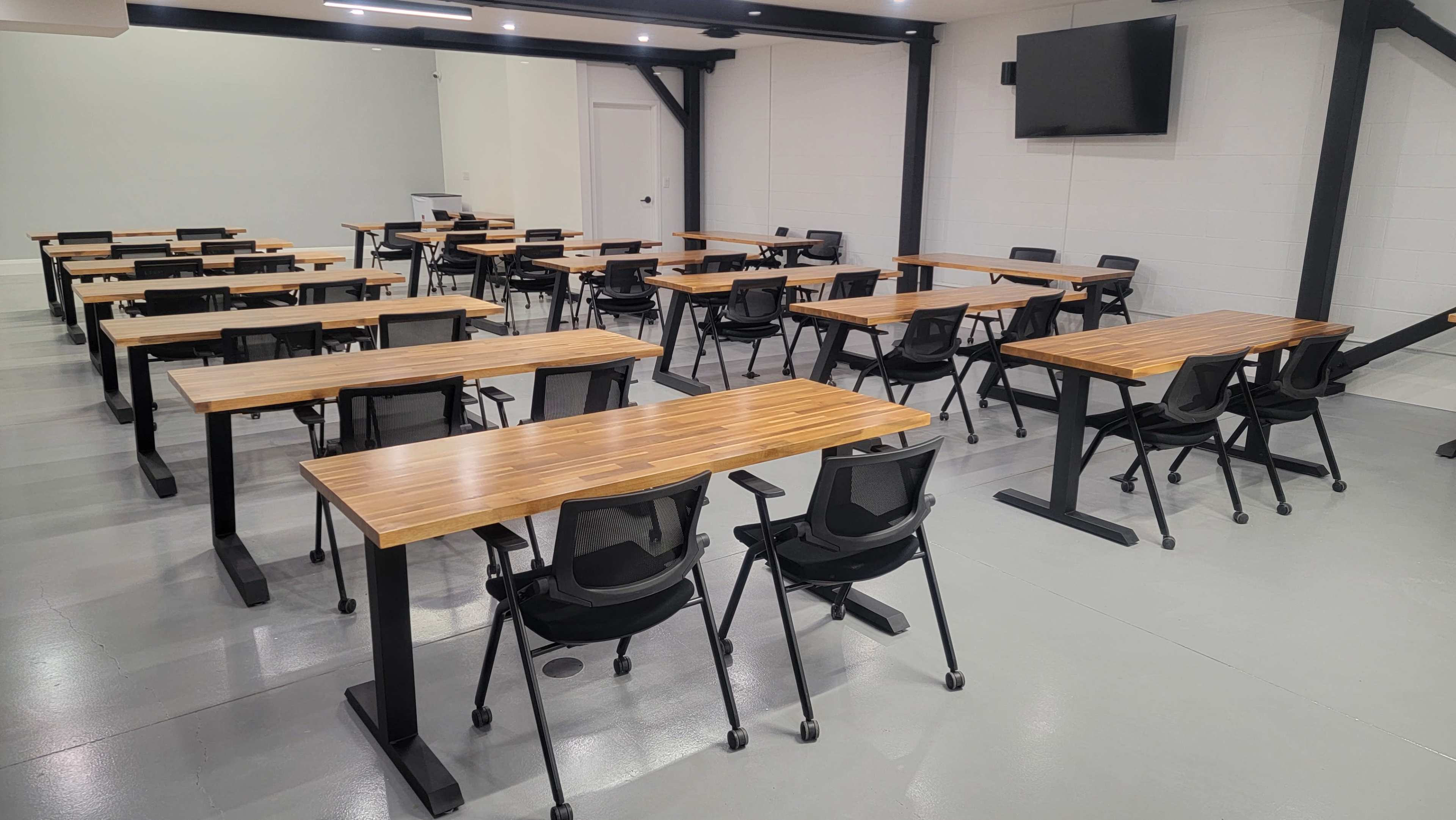 The image shows a classroom or meeting room with multiple rows of wooden tables and rolling chairs arranged in a spacious area.