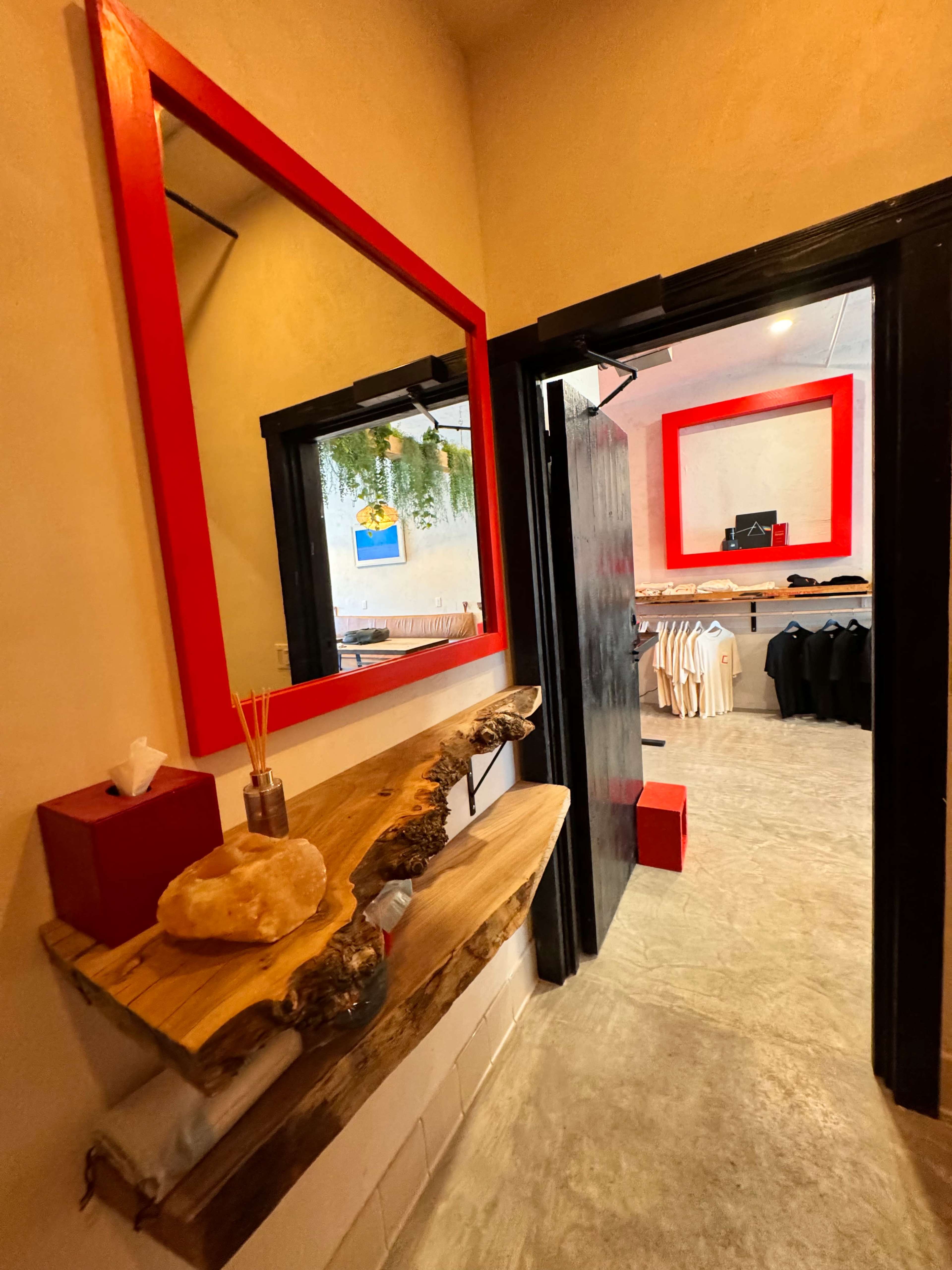 A small room with a wooden shelf, a red-framed mirror, and black clothing displayed on hangers inside.