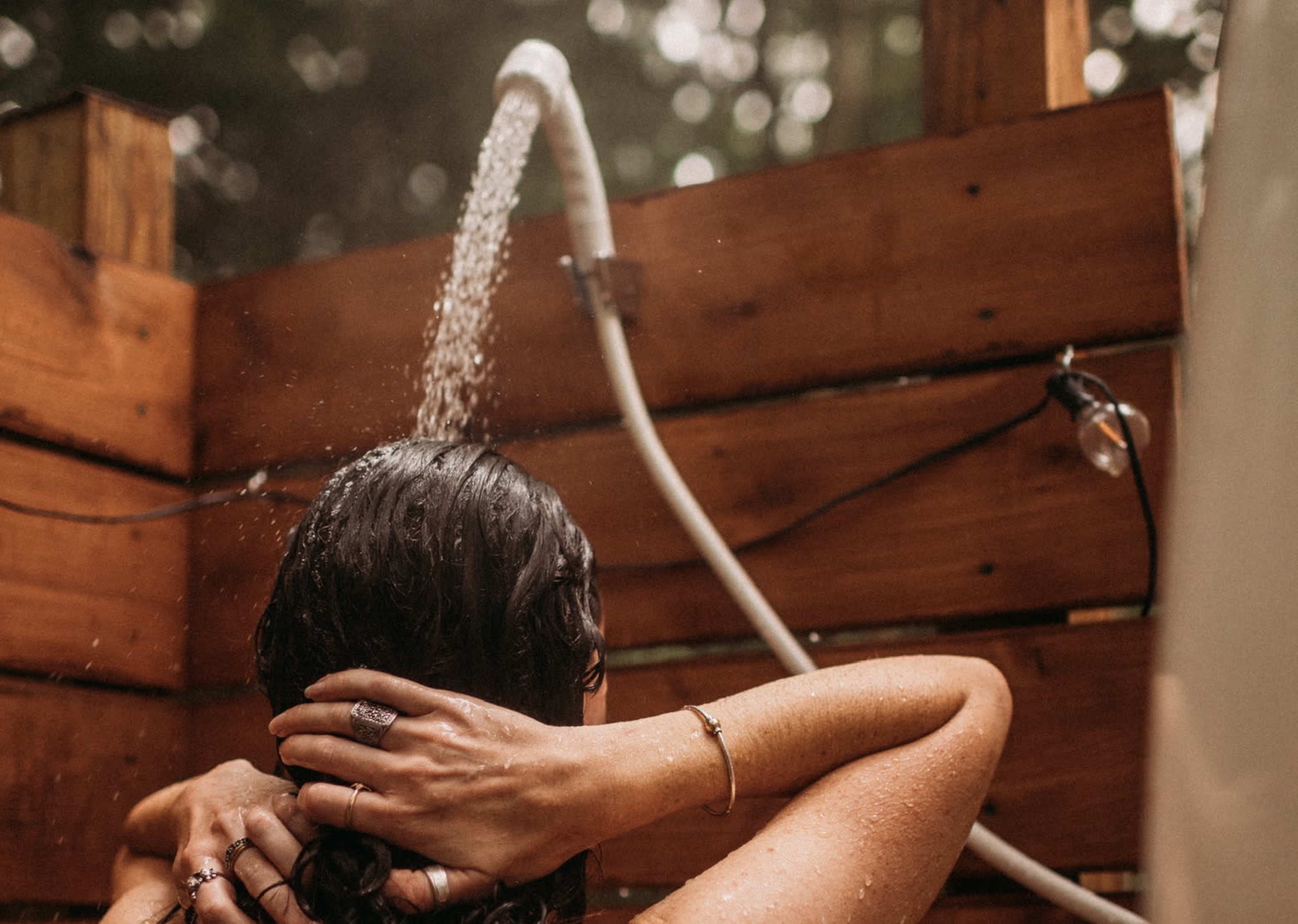 A person with wet hair stands under a showerhead in an outdoor setting surrounded by wooden walls.