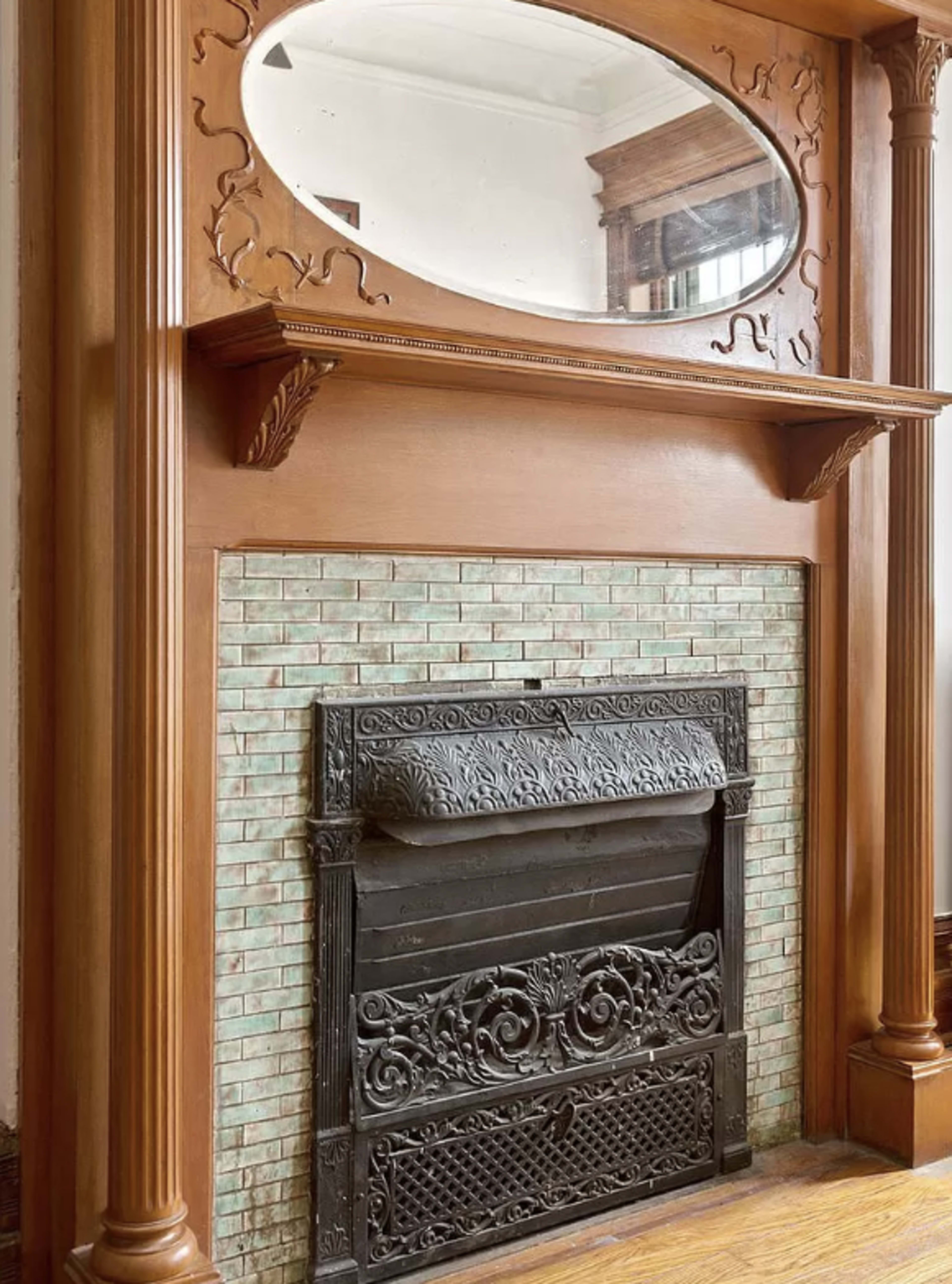 The image shows an ornate wooden fireplace mantel with a large oval mirror above it and an intricately designed black iron fireplace insert, surrounded by green tiles.