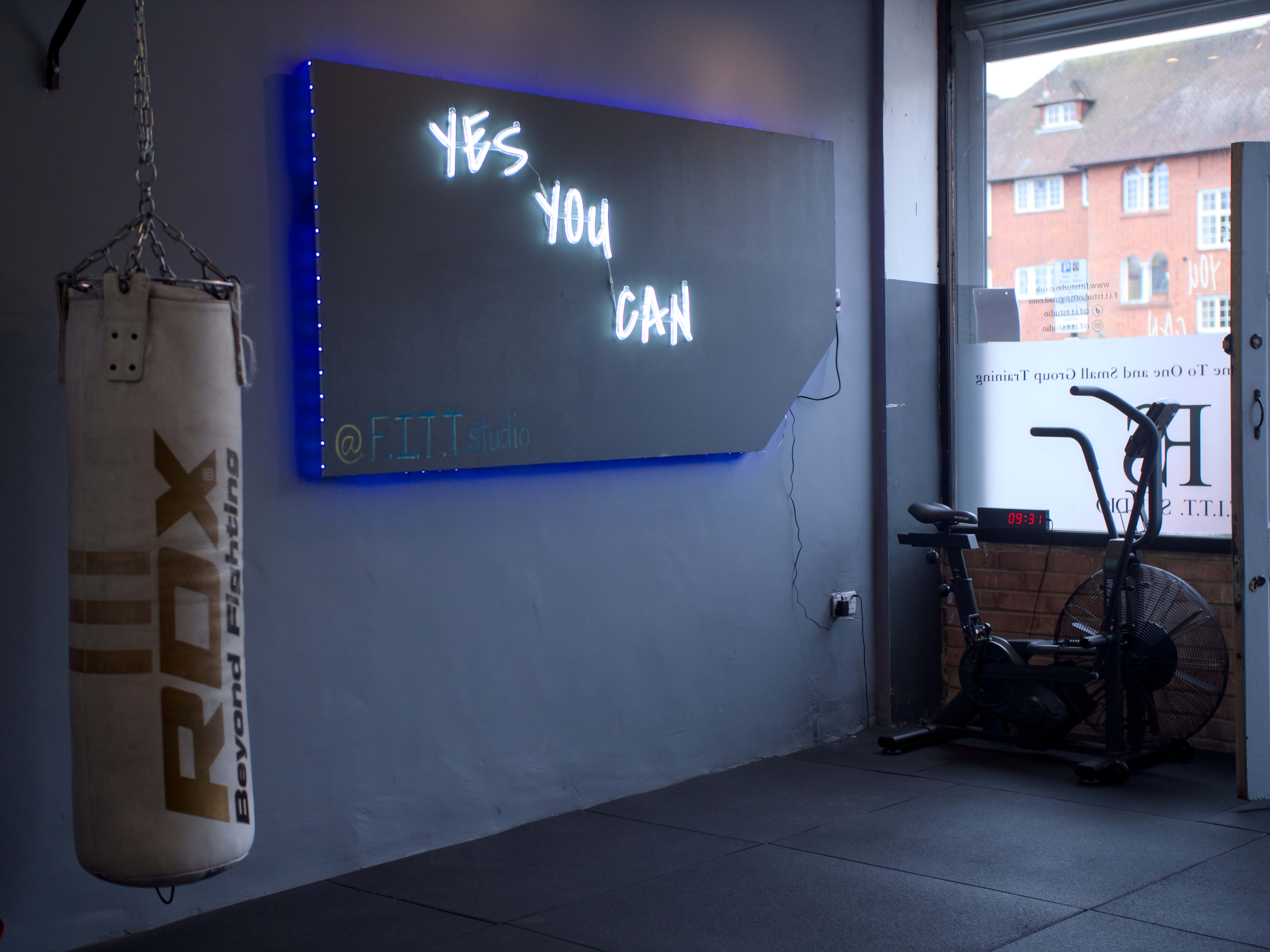 The image shows an interior space of a gym featuring a punching bag, an exercise bike, and a wall-mounted sign that reads "YES YOU CAN."