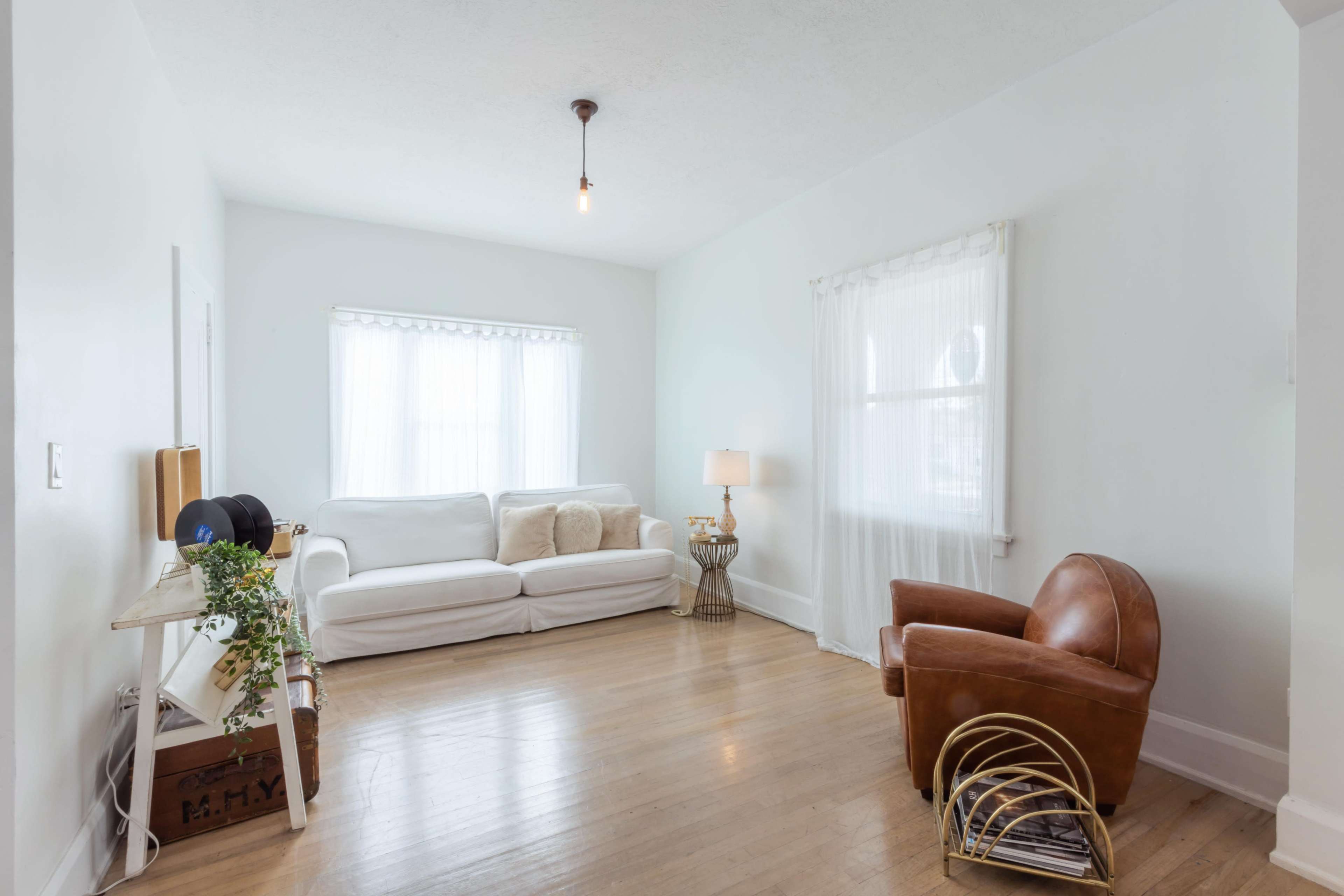 The room features a white sofa, a wooden side table with a lamp, and a brown leather armchair, all set against light-colored walls and hardwood flooring.