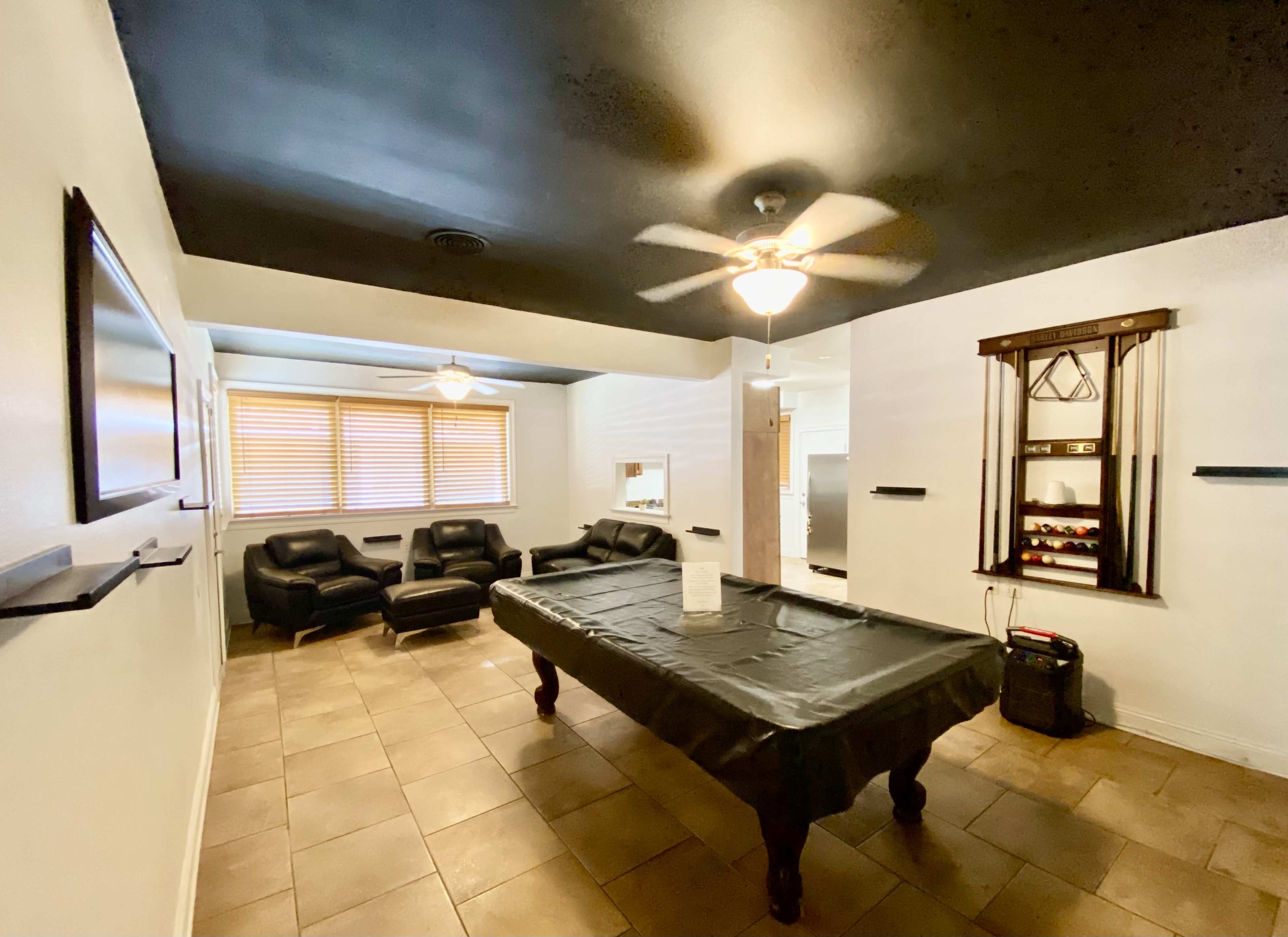 The image shows a game room featuring a pool table, black leather seating, and ceiling fans under a black ceiling.
