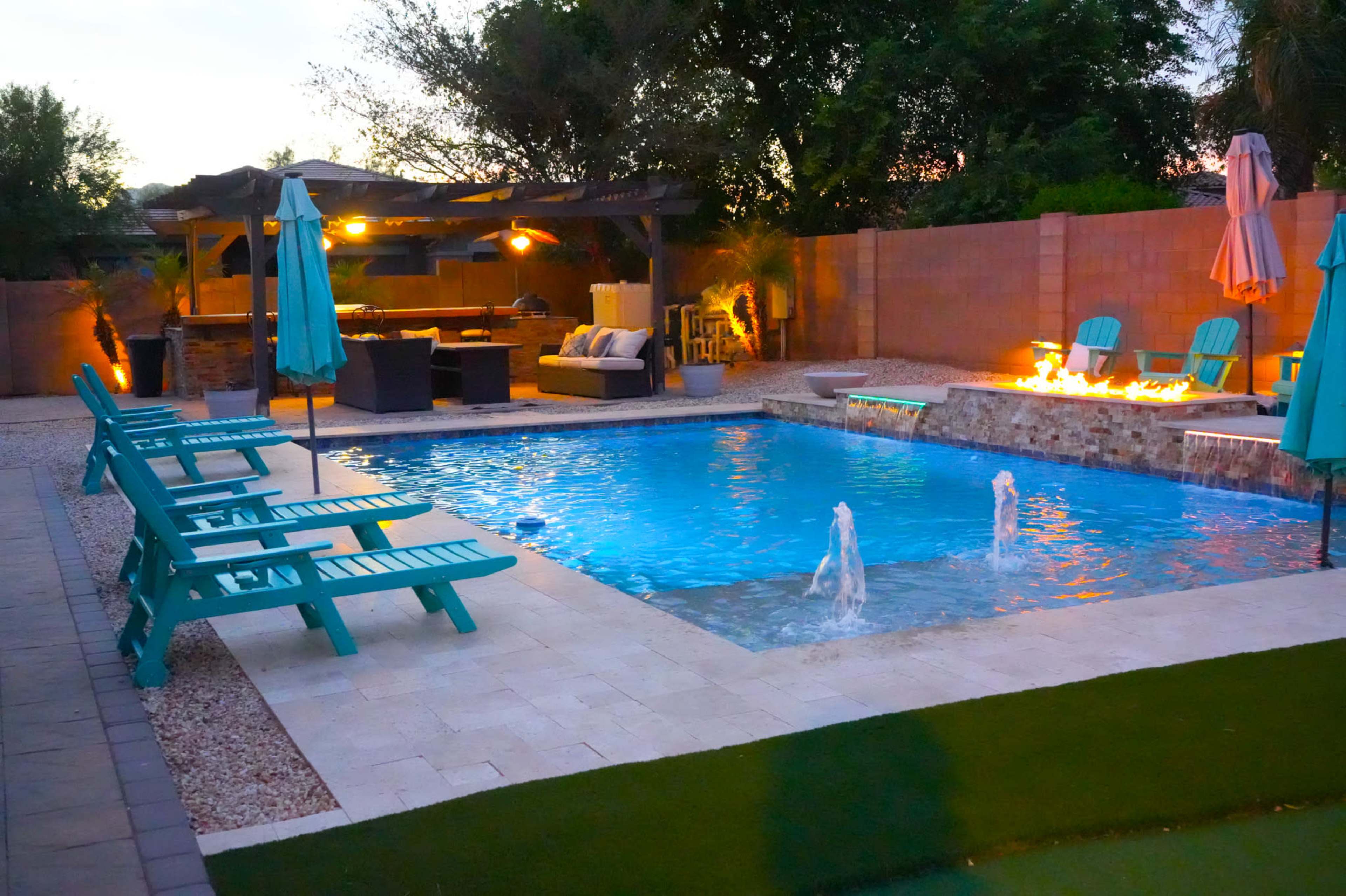 The image shows a backyard pool area with turquoise lounge chairs, a water feature, and a seating area under a pergola, surrounded by trees and illuminated by outdoor lighting.