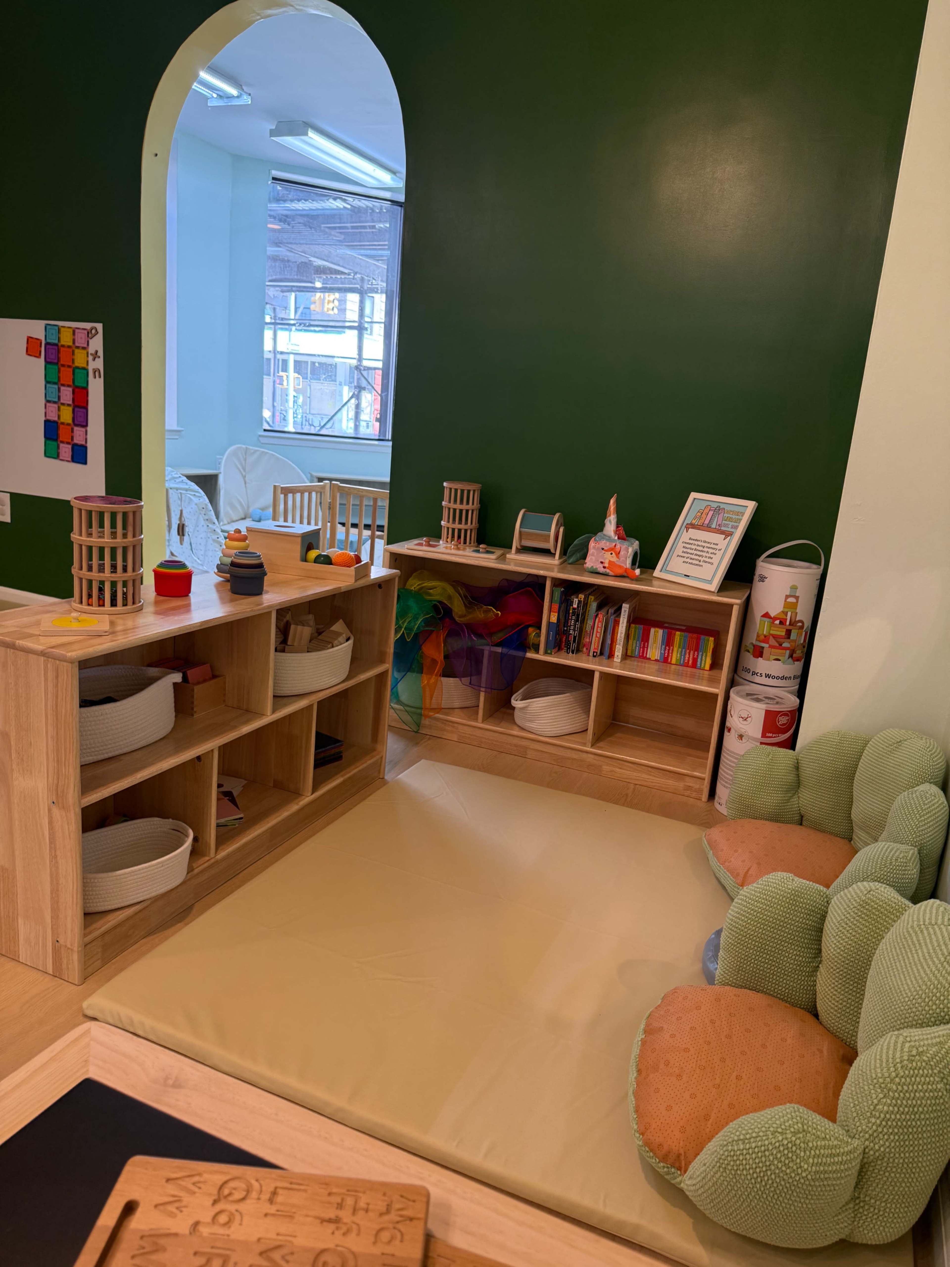 The image shows a colorful children's play area featuring wooden shelves with toys, a soft mat on the floor, and a green wall in the background.