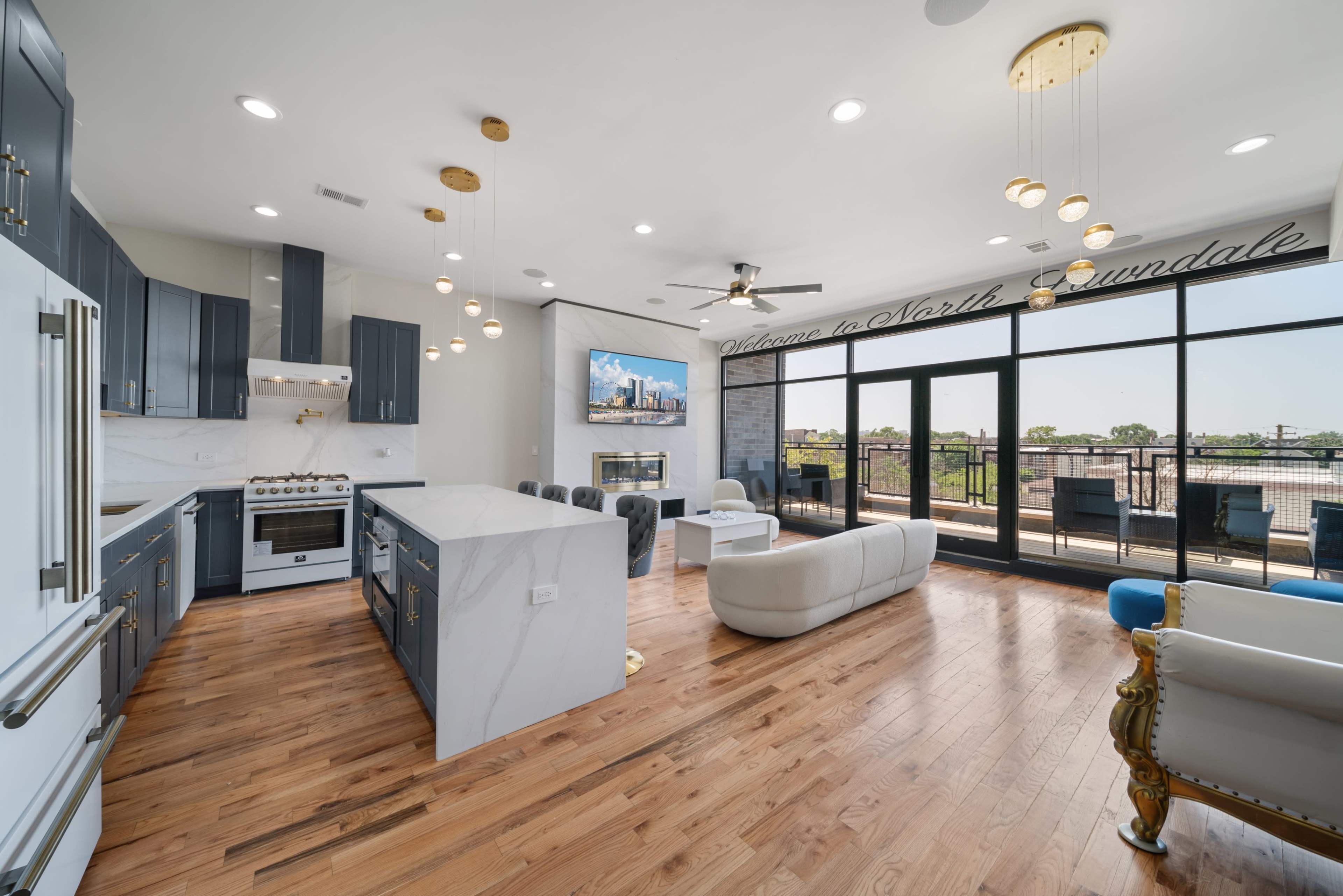 The image shows a modern kitchen and living space with blue cabinetry, wooden flooring, and large windows overlooking a balcony.