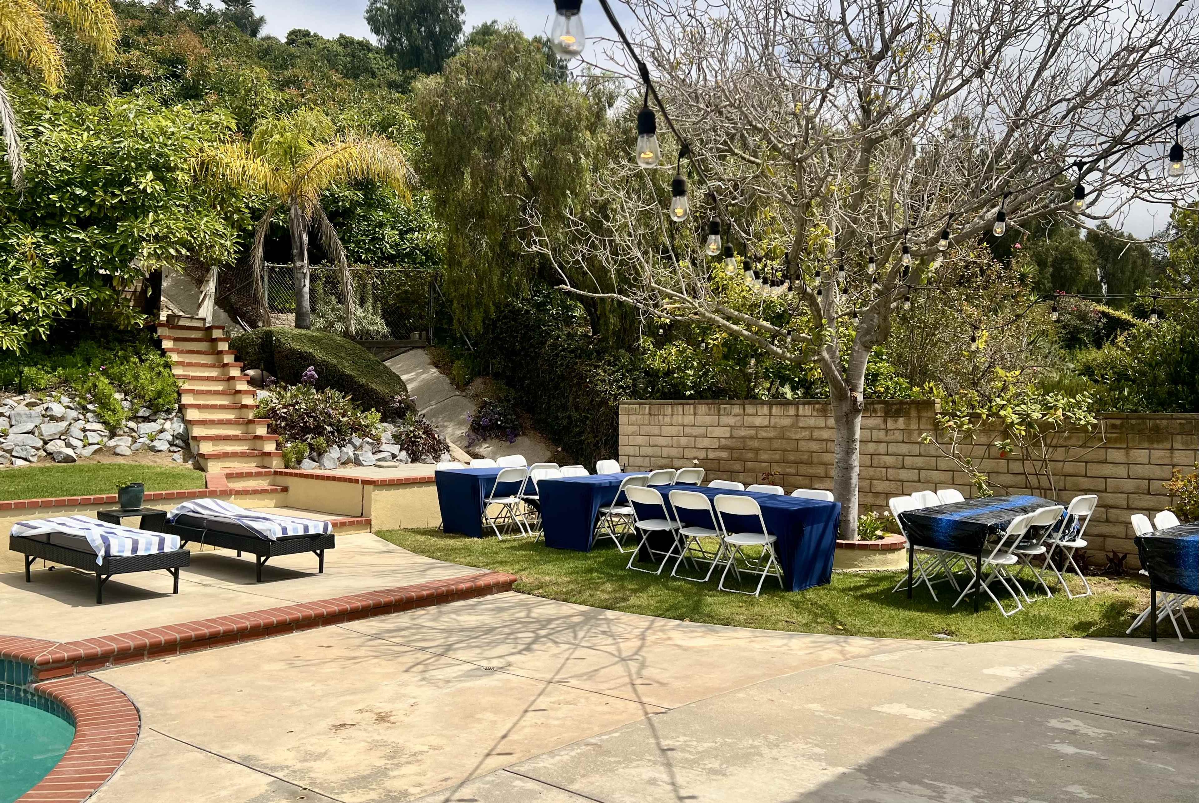 The image shows a backyard setting with a swimming pool, several lounge chairs, and tables set up for an outdoor event, surrounded by lush greenery and a paved pathway.
