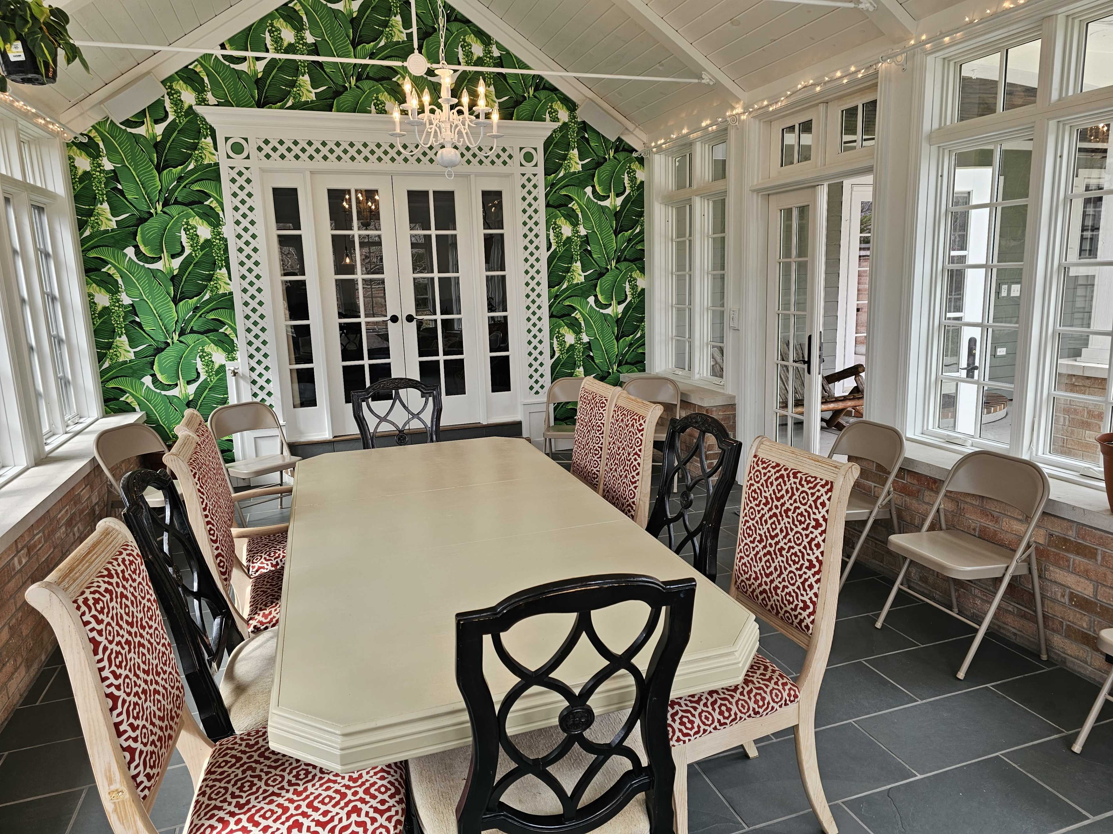 A bright sunroom features a large table surrounded by eight ornate chairs, with a leafy green wallpaper and glass doors leading outside.