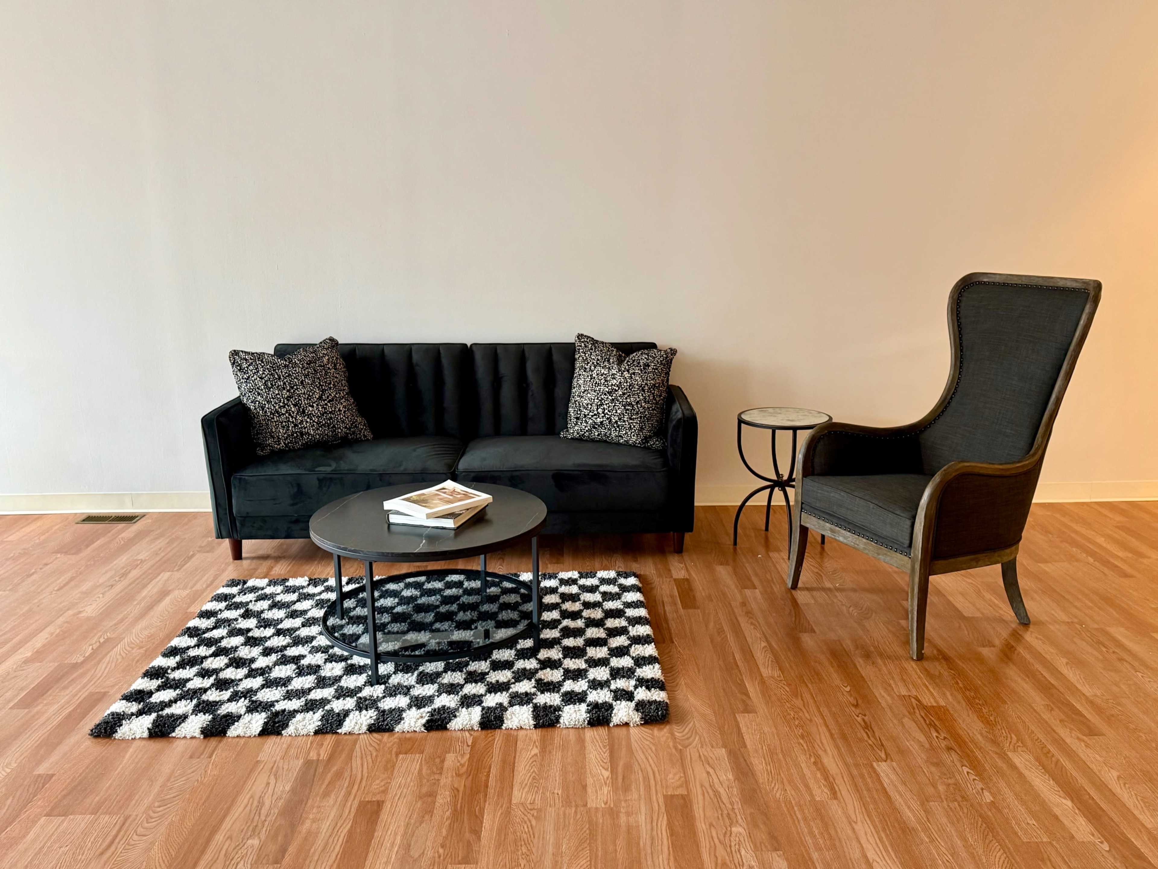 A black sofa with patterned cushions sits opposite a tall-backed chair beside a small round table on a checkered rug.