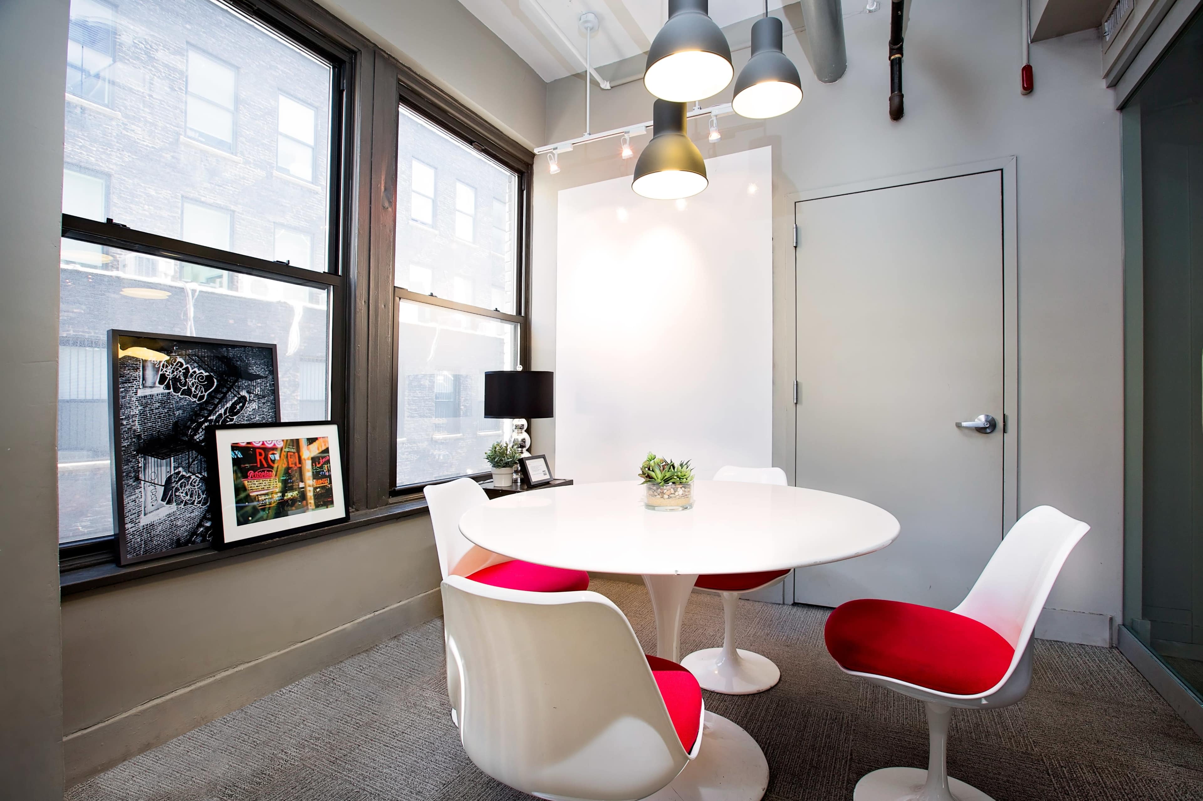 A modern dining area features a round white table surrounded by four white and red chairs, with large windows and a plant centerpiece.