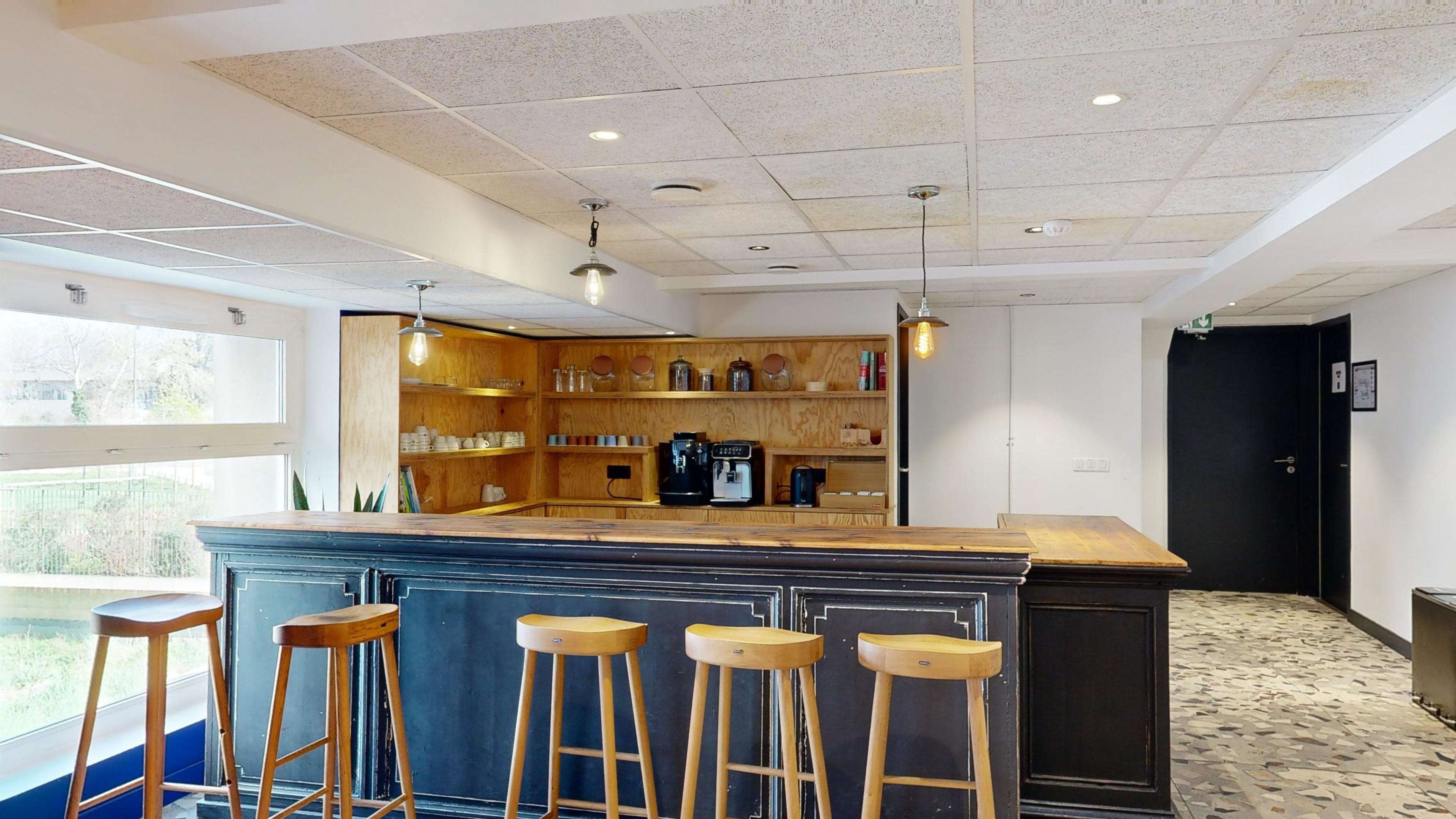 A modern bar area with a wooden counter and four high stools, set against a backdrop of shelves displaying various containers and a coffee machine.