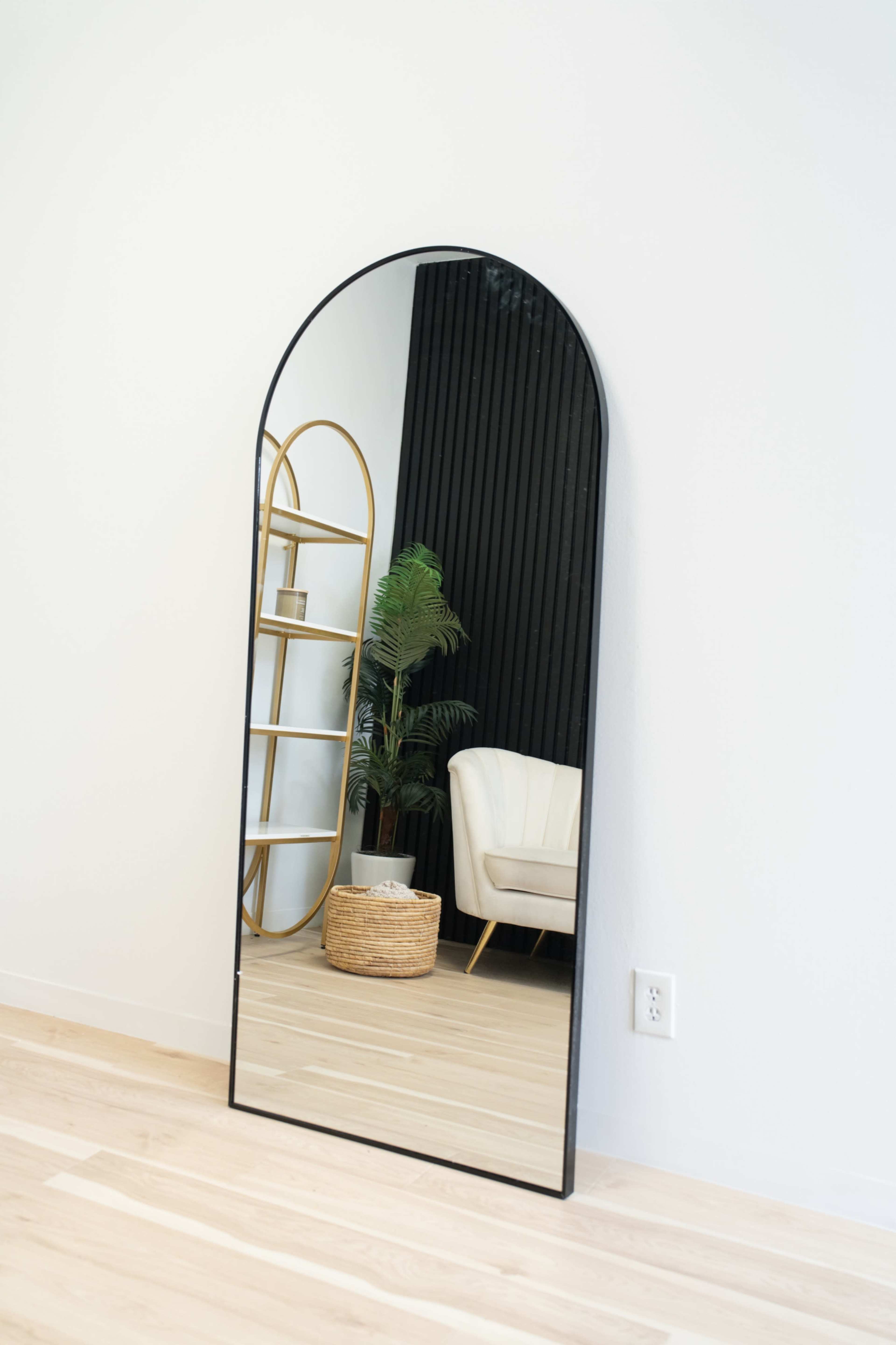 A large, arched mirror leans against a white wall, reflecting a chair and a gold-framed shelving unit beside a potted plant.