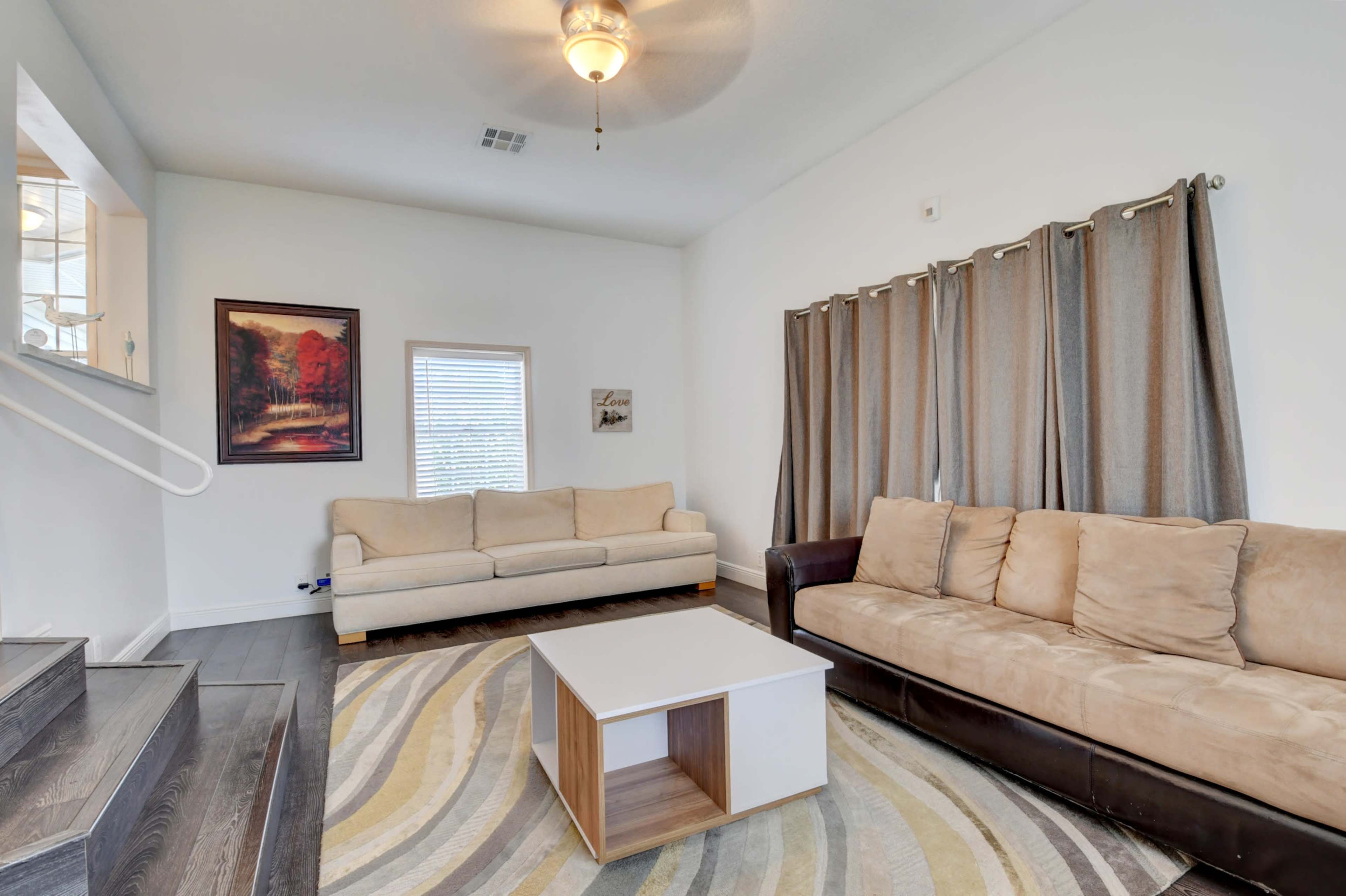 The living room features a beige sectional sofa, a brown couch, a light-colored coffee table, and large windows covered with gray curtains.