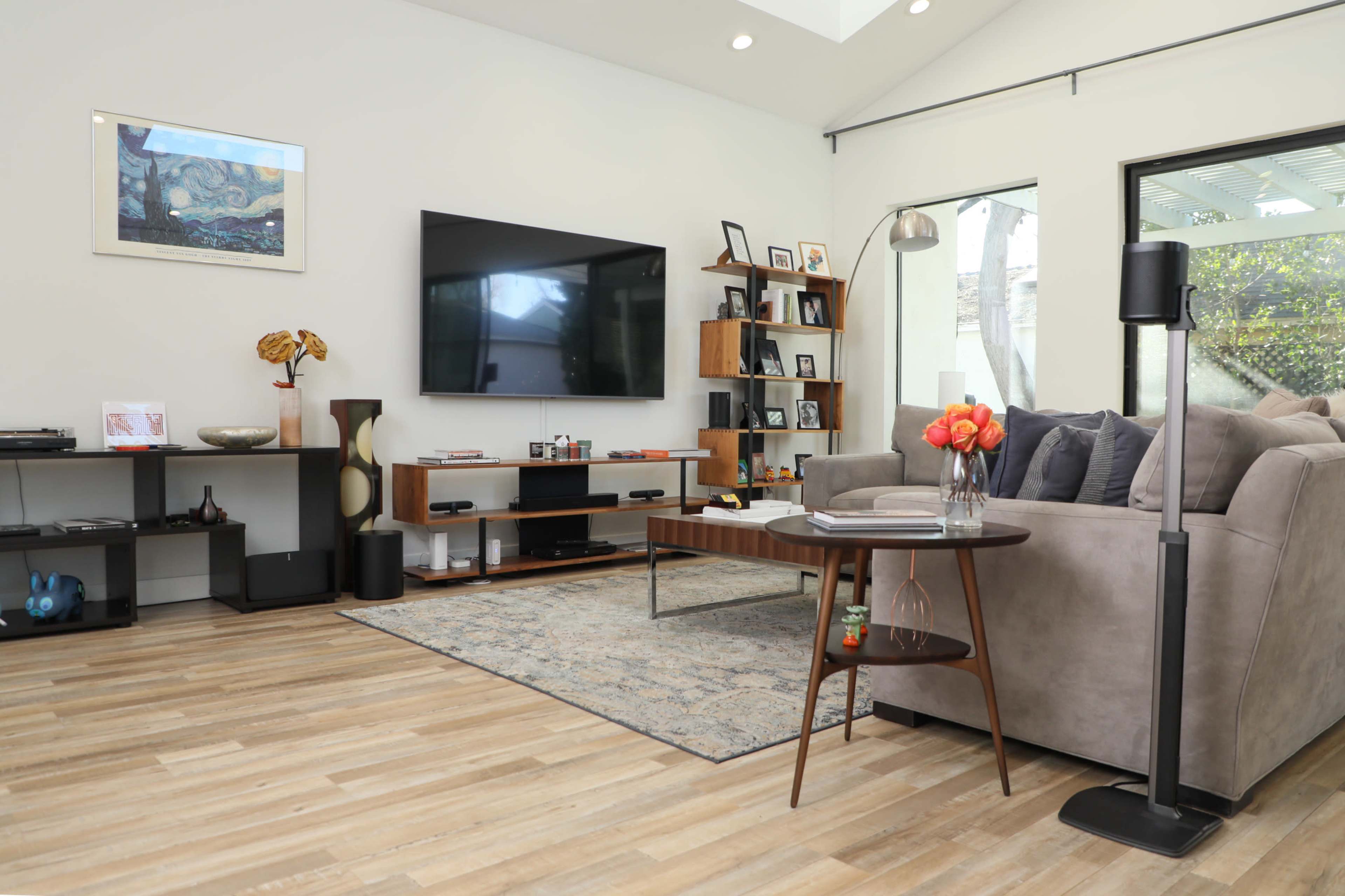 The living room features a modern sofa, a coffee table with flowers, a large television mounted on the wall, and a mix of shelves and decorative items.