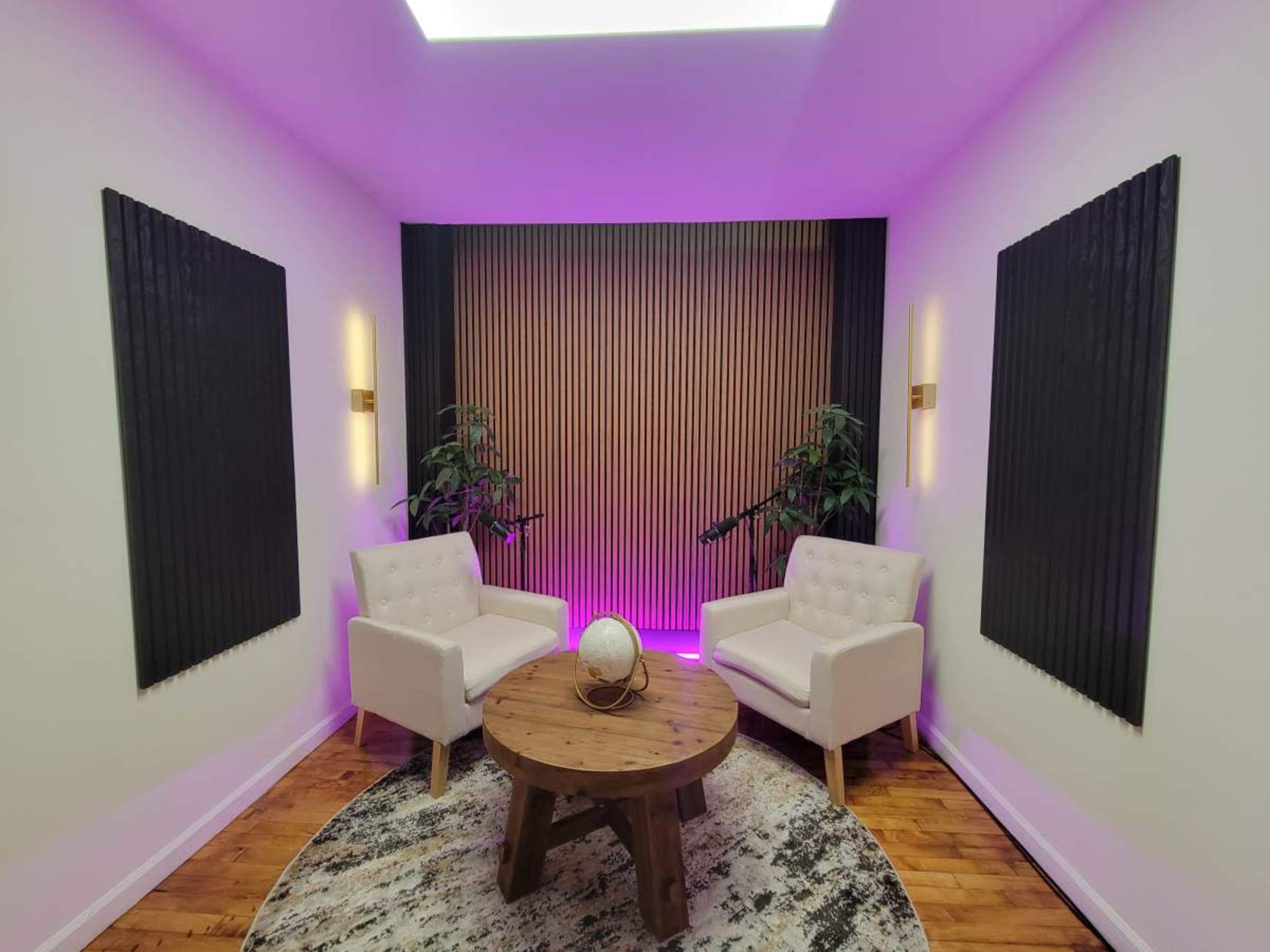 The image shows a modern, minimalist room with two white chairs, a round wooden table, and decorative plants, all illuminated by soft purple lighting.