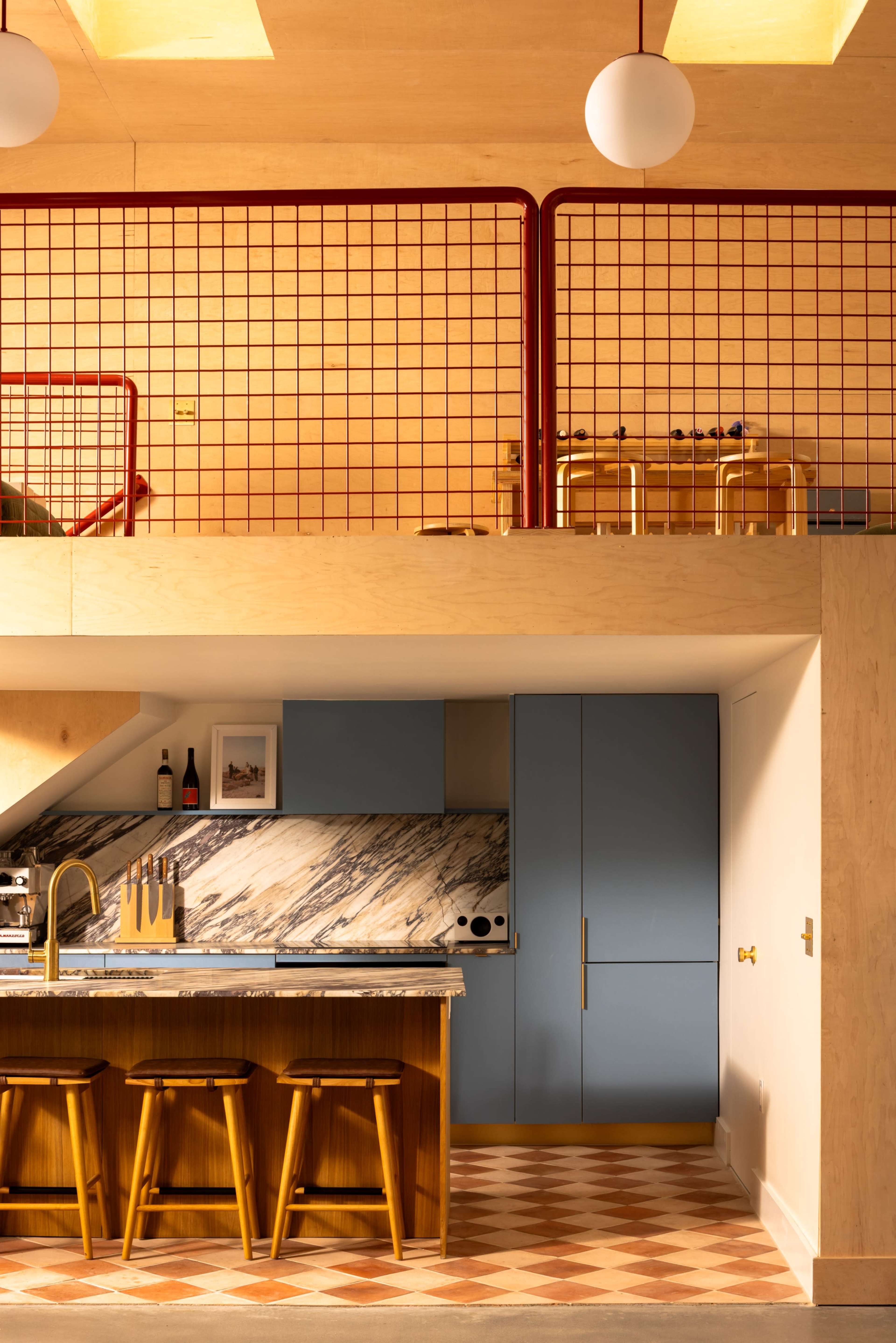 The image shows a modern kitchen with a marble countertop, wooden bar stools, and a blue refrigerator, situated beneath a lofted sleeping area with a red metal railing.