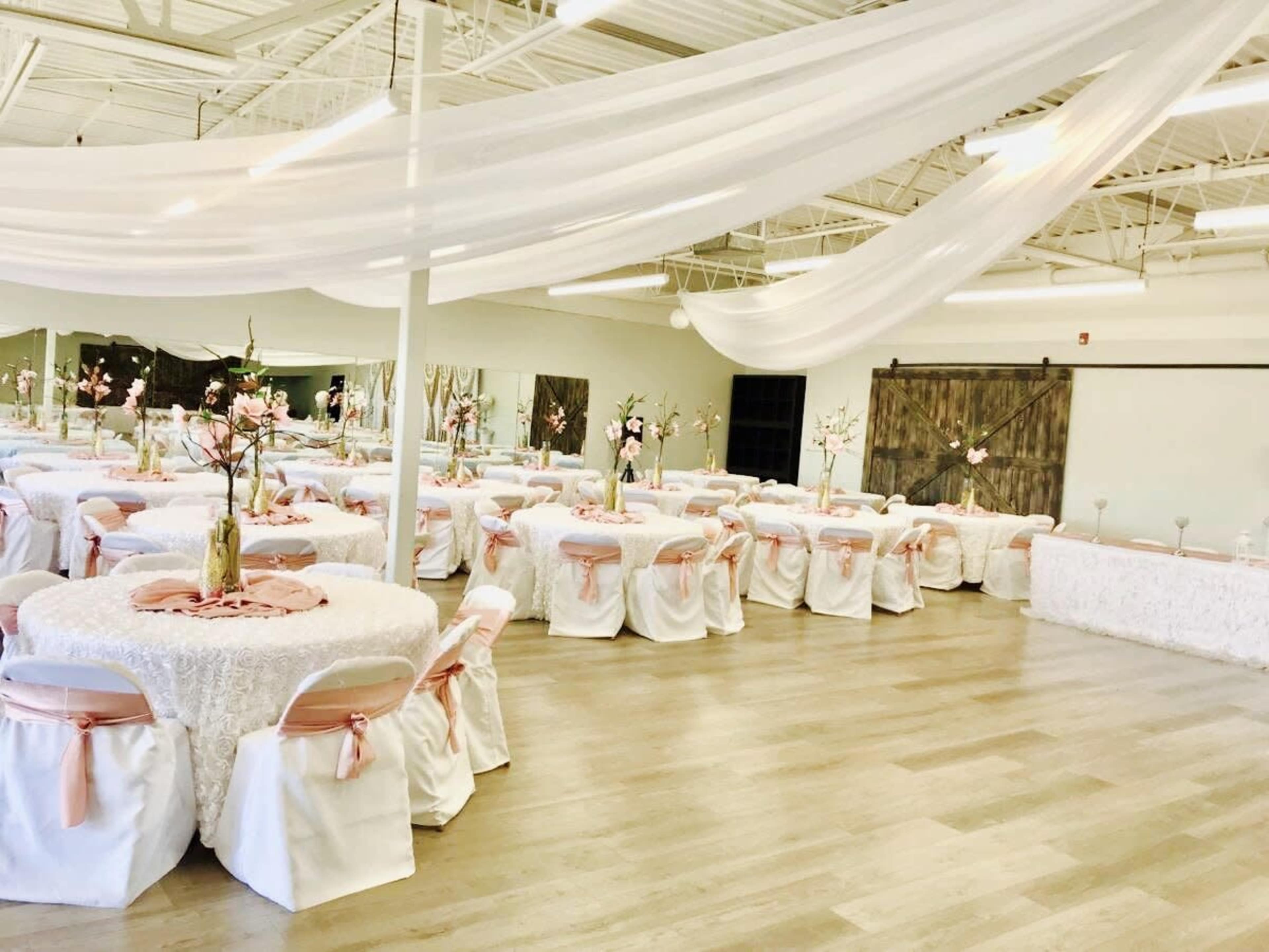 A spacious event hall is set up for a banquet, featuring round tables dressed in white linens with pink sashes and floral centerpieces.