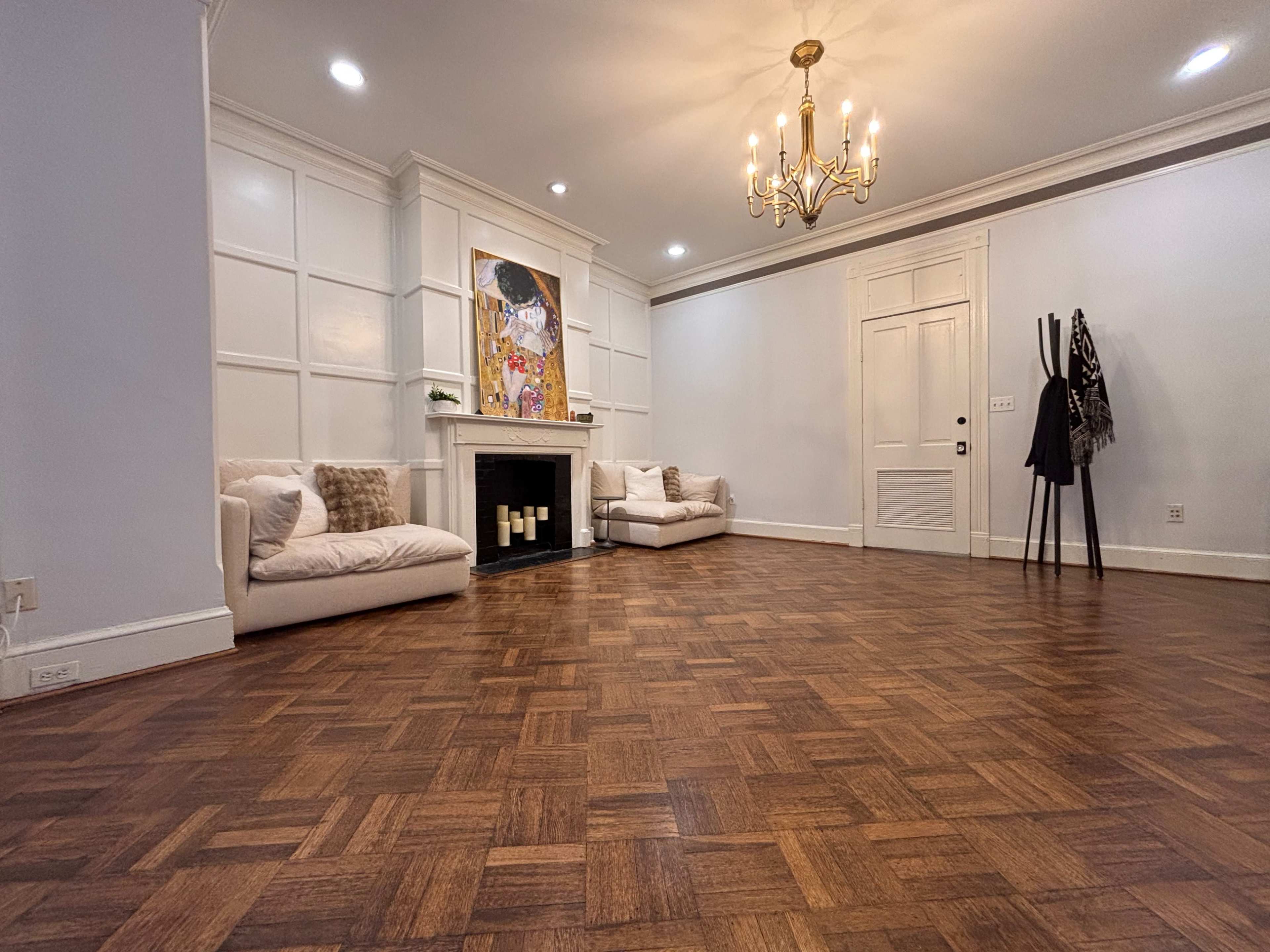 The room features hardwood flooring, an ornate chandelier, a fireplace, and two white couches against the walls.
