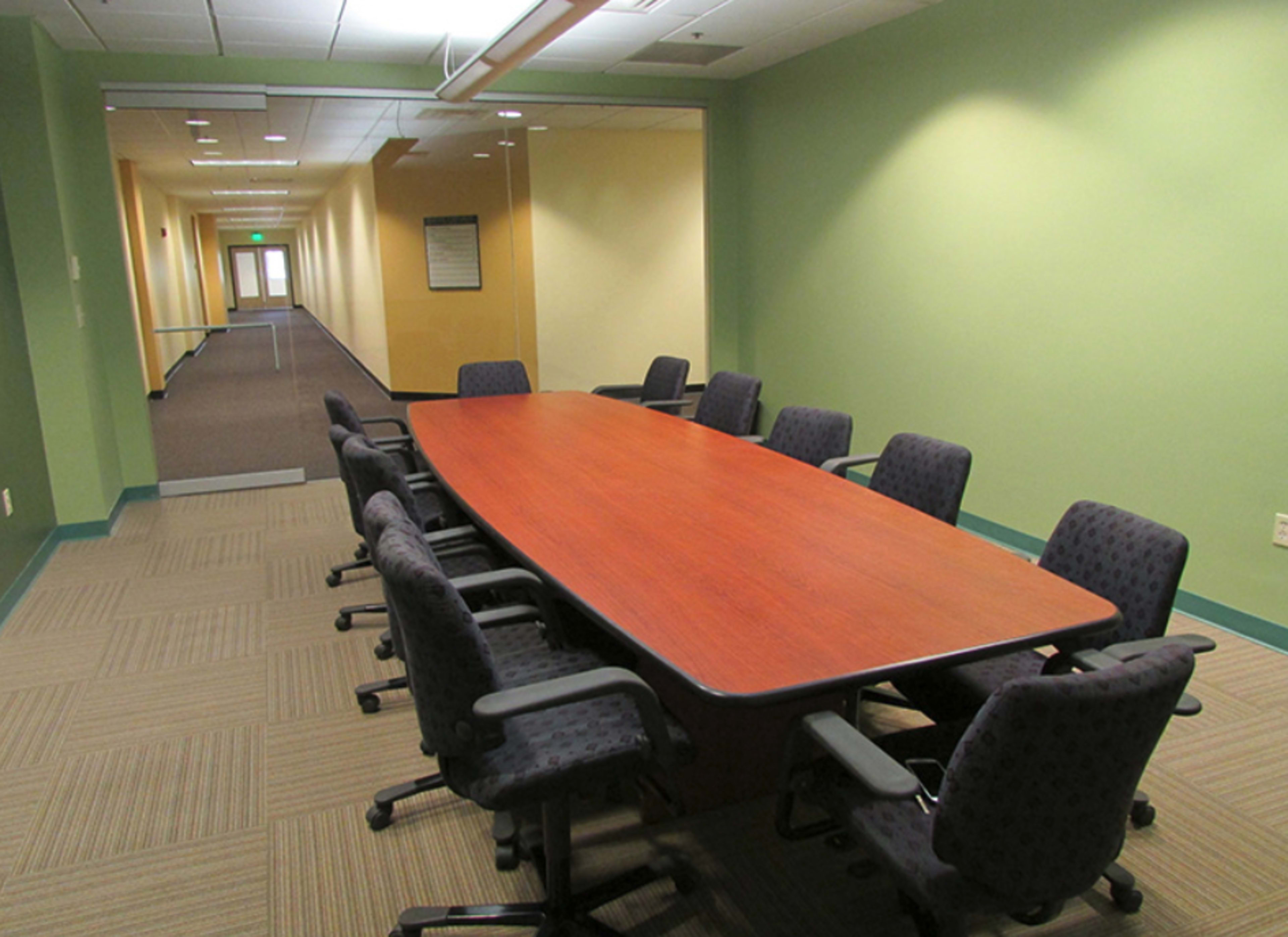 A long, wooden conference table is surrounded by eight upholstered chairs in a brightly lit office space.