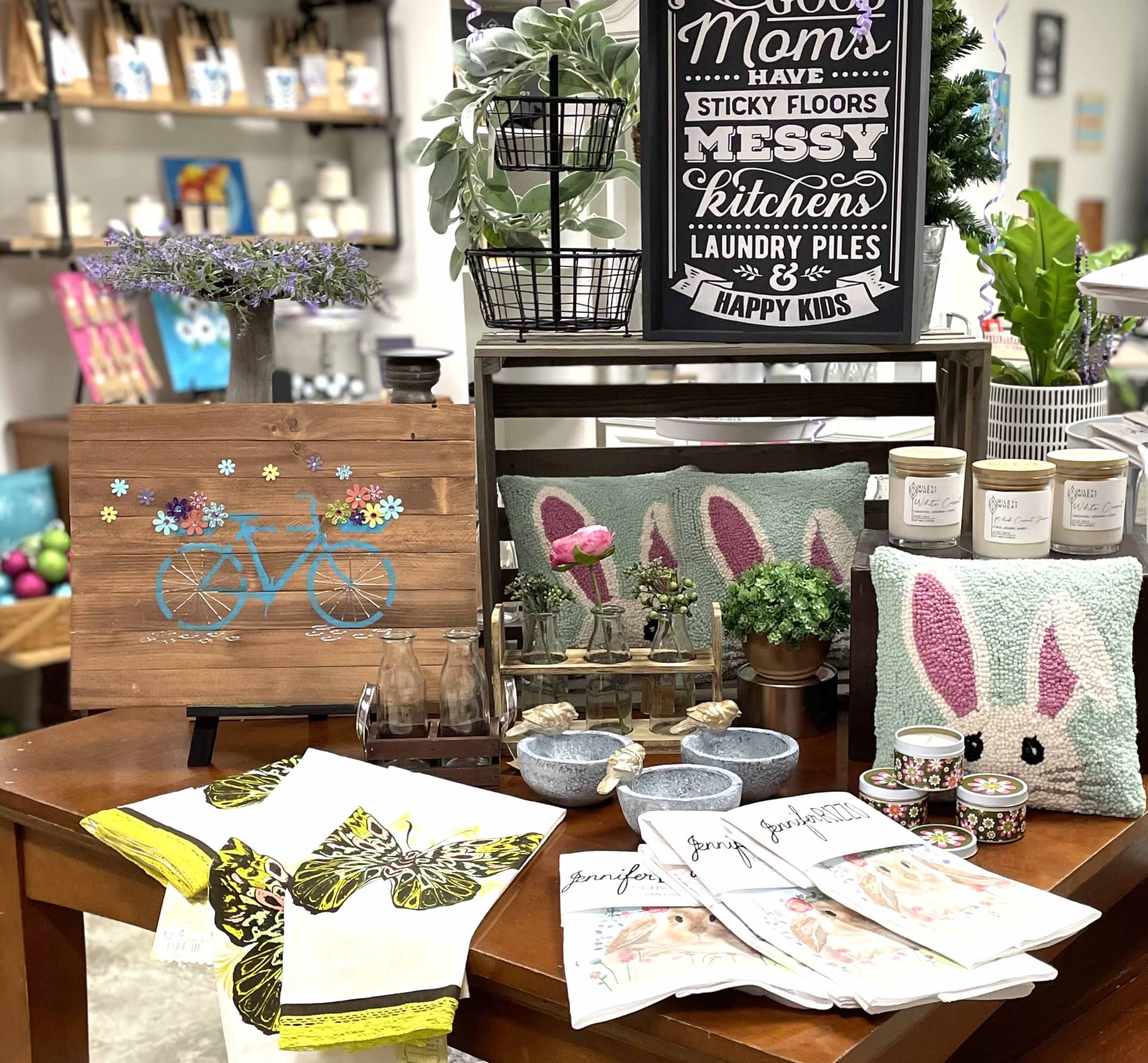 The image features a table display with decorative items, including a wooden sign, throw pillows shaped like bunnies, kitchen towels, and various containers with plants and stationery.