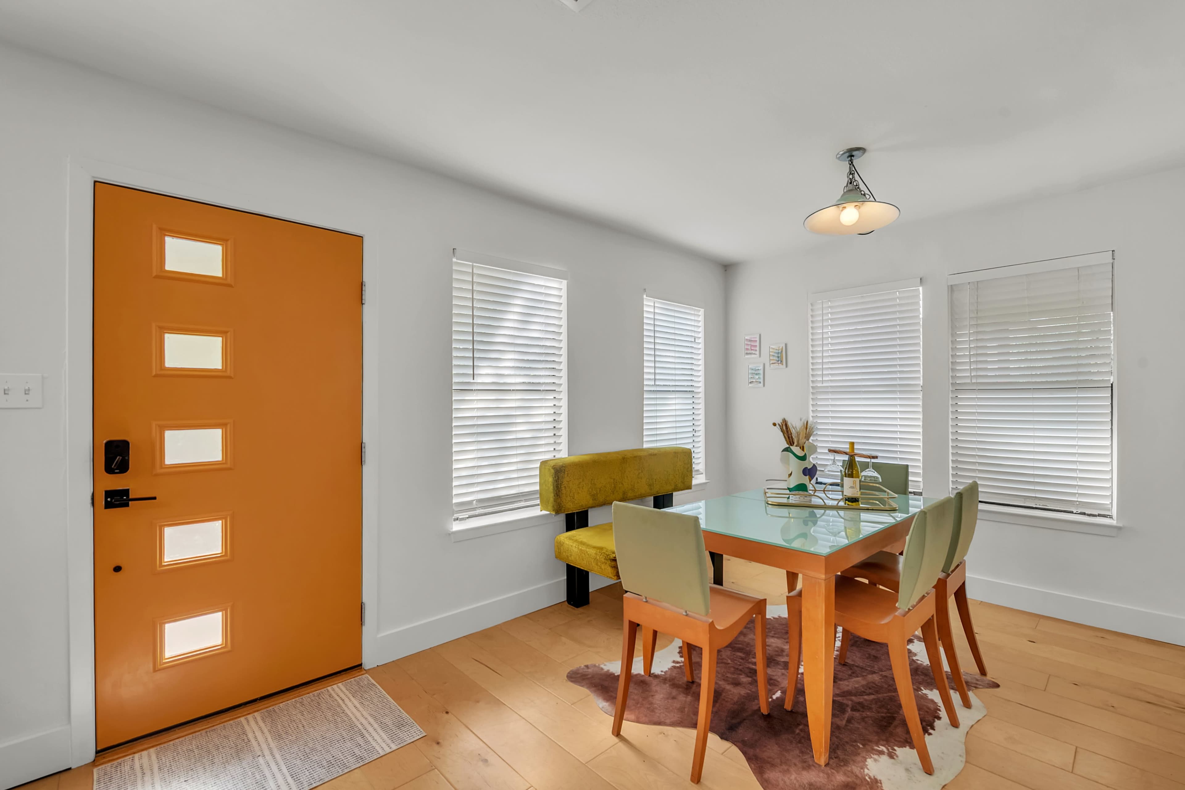 The image features a bright dining area with an orange front door, a glass table surrounded by four chairs, and a yellow upholstered bench against the wall.