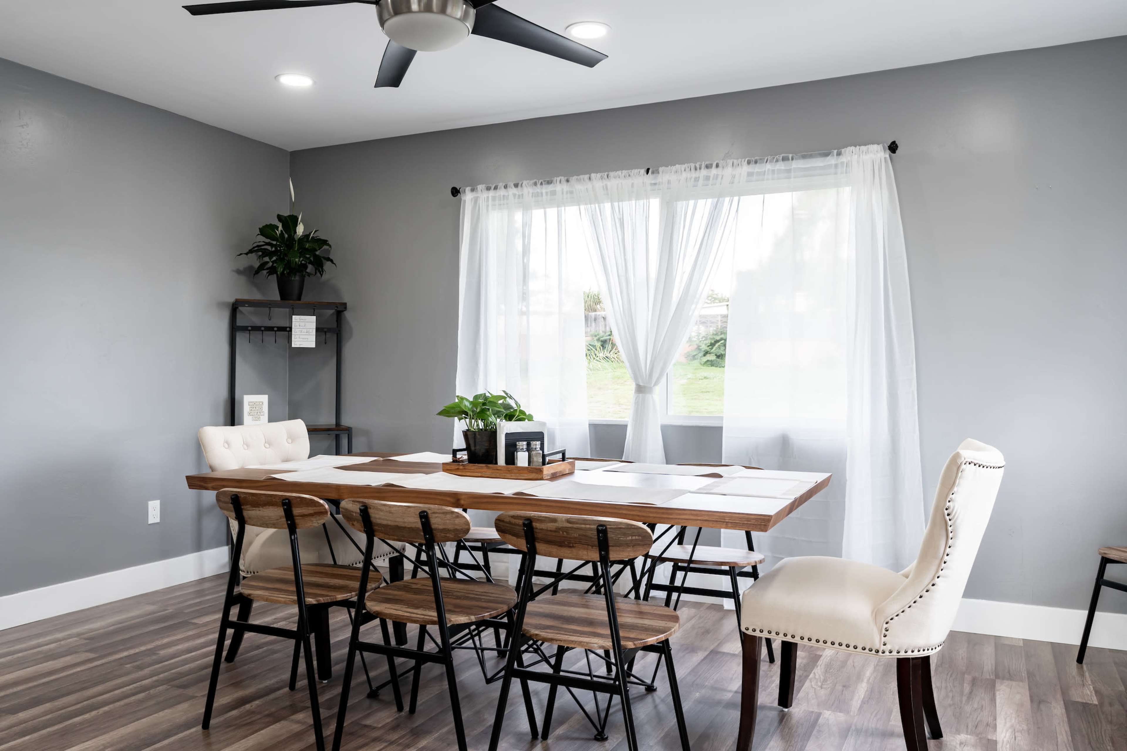 A modern dining area features a wooden table surrounded by chairs, with a large window dressed in sheer curtains allowing natural light to enter.