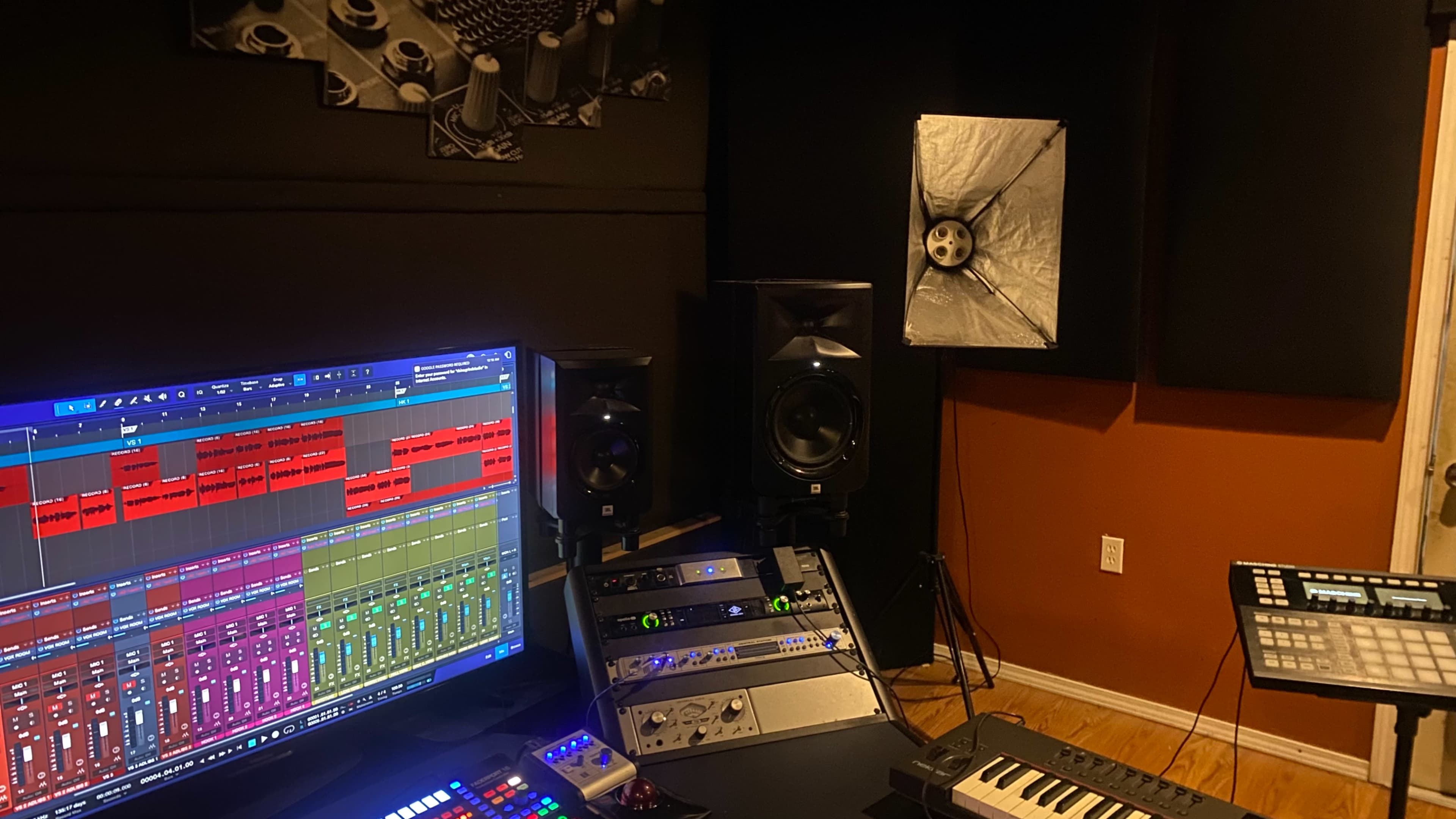 A recording studio setup features a large computer monitor displaying audio tracks, mixing equipment, studio speakers, and a keyboard.