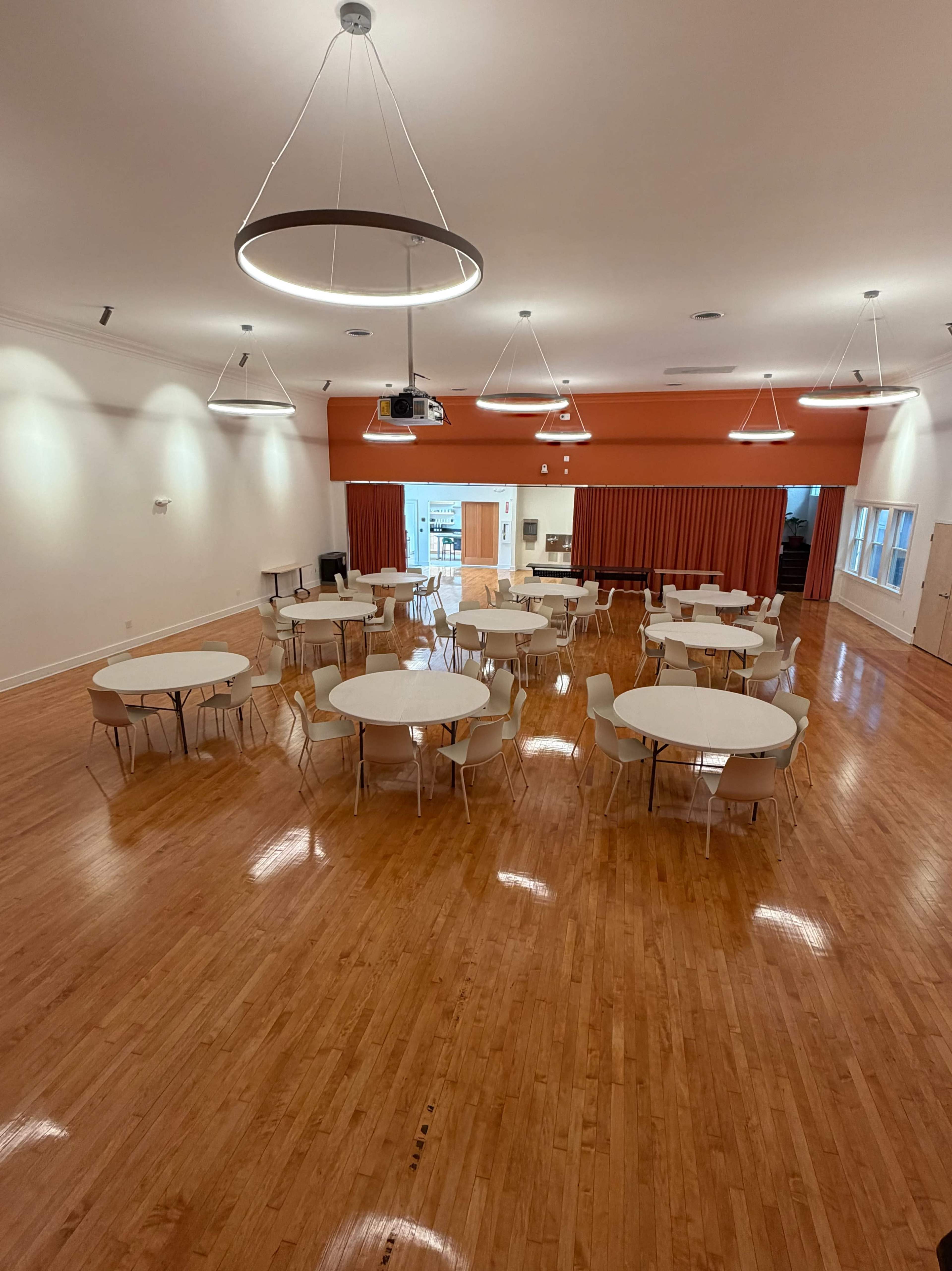 The image shows a spacious indoor hall with several round tables and chairs arranged neatly on polished wooden flooring under modern lighting fixtures.