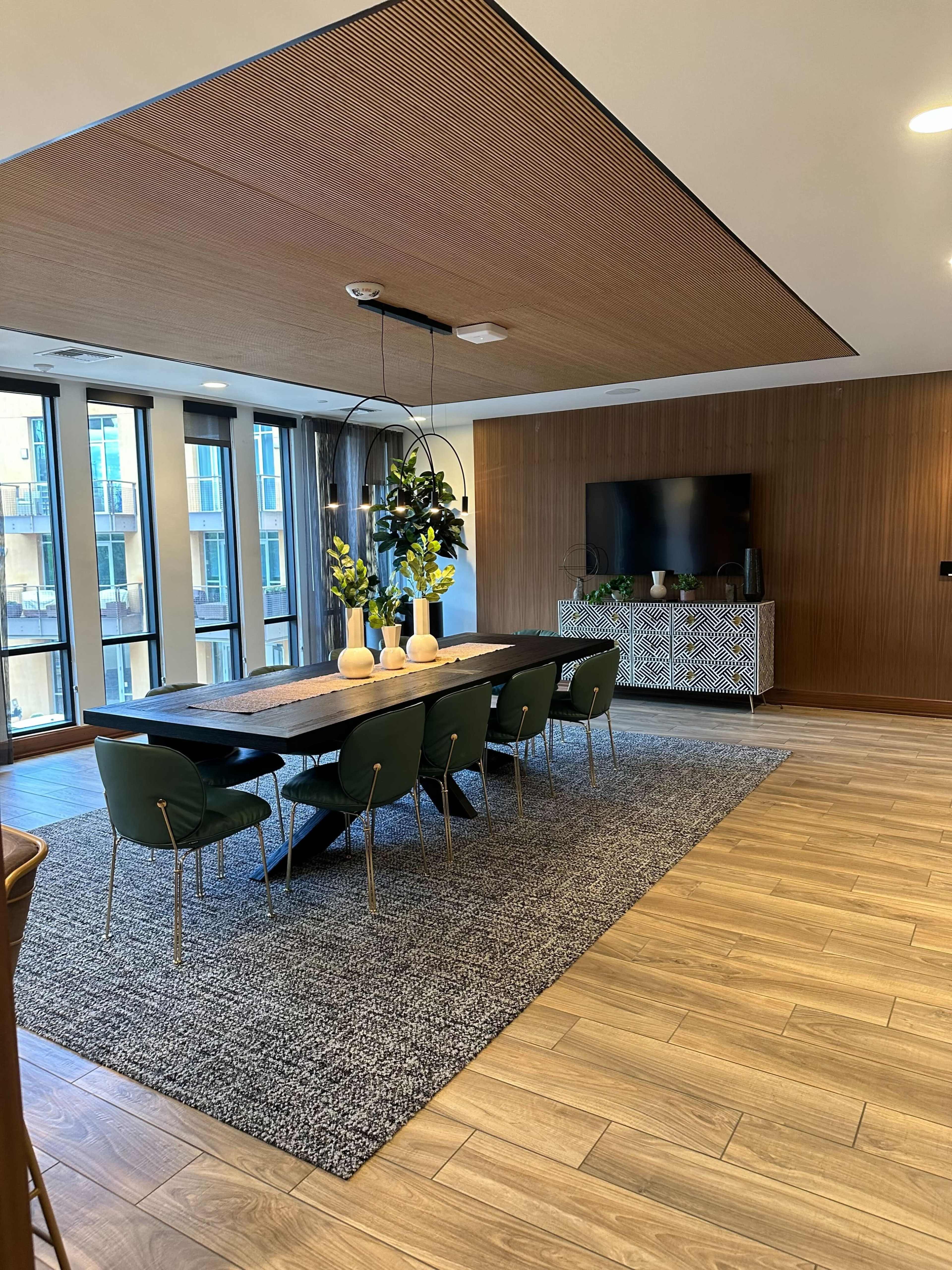 A modern dining area with a large table surrounded by green chairs, a wall-mounted TV, and decor elements like a rug and plants.