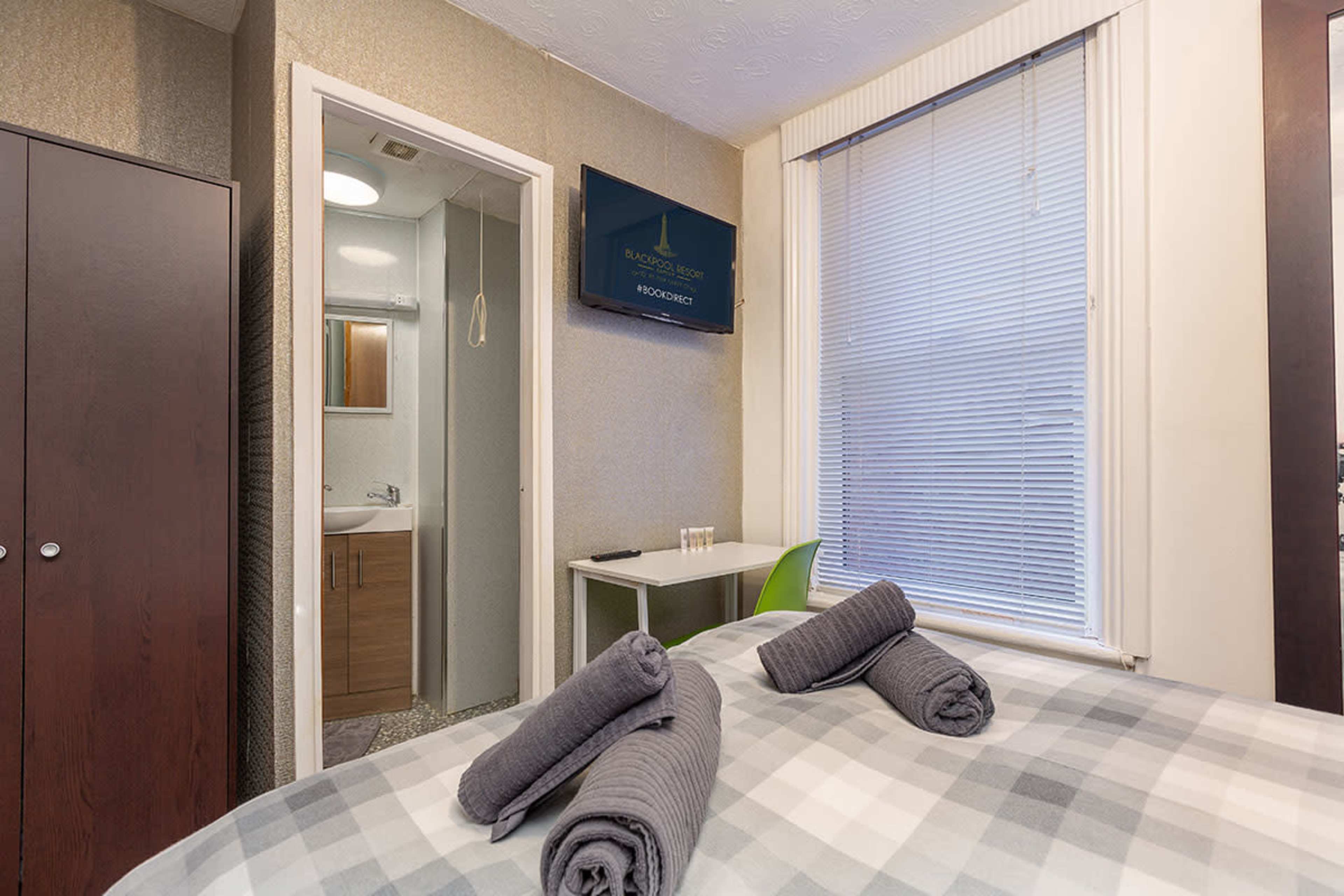 A neatly arranged bedroom with a double bed, a TV mounted on the wall, a small table with a chair, and a visible en-suite bathroom area.